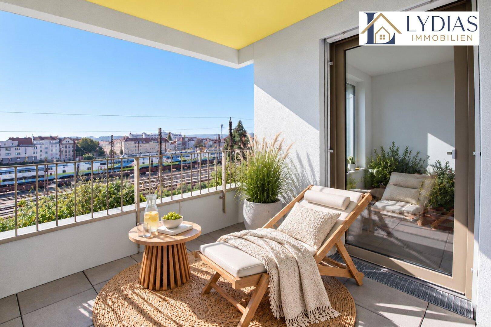 Sonniger Balkon mit Liegestuhl, Pflanzen und Blick auf die Bahngleise und Stadt.
