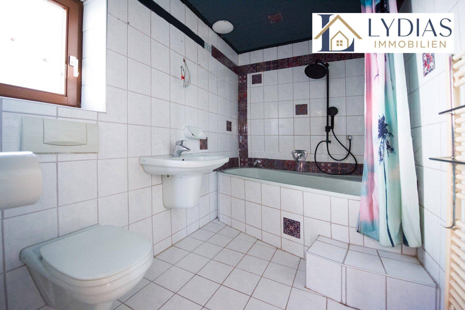 Gefliestes Badezimmer mit Badewanne, Dusche und Fenster, in älterem Zustand.