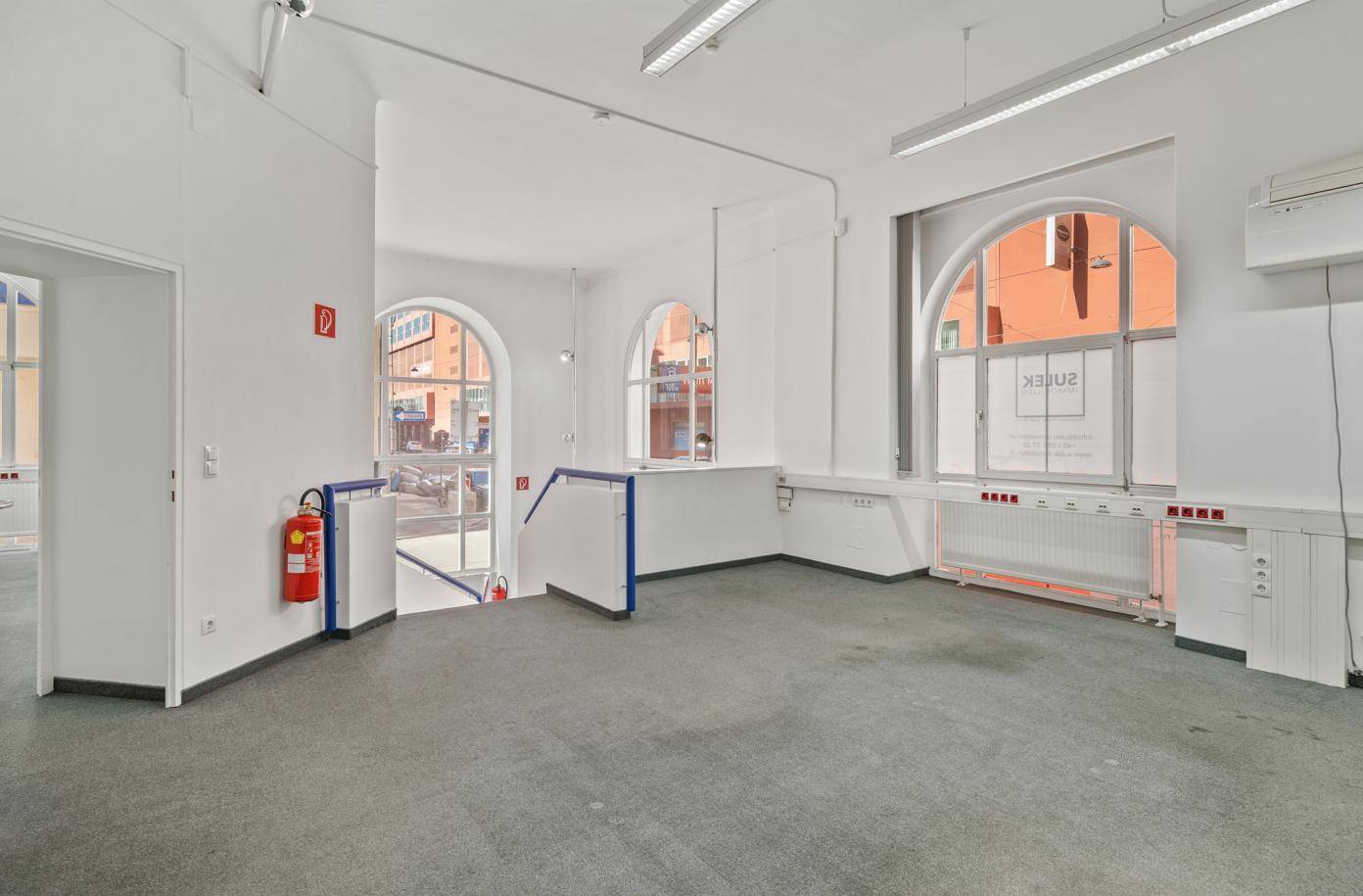 Heller Büroraum mit Teppichboden, großen Bogenfenstern und Zugang zu einem weiteren Bereich.