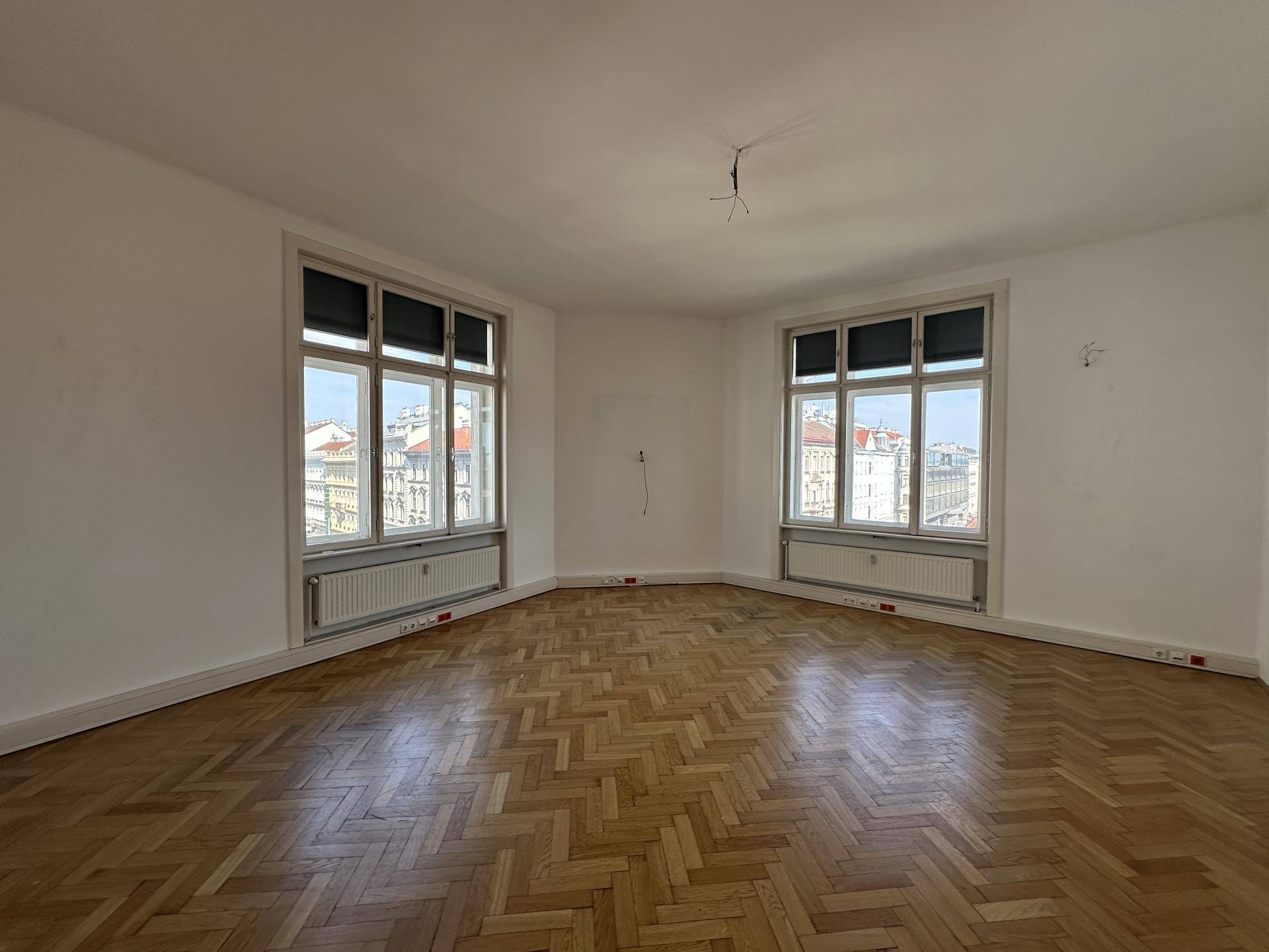 Geräumiger, heller Raum mit Fischgrätparkett und zwei großen Fenstern mit Stadtblick.