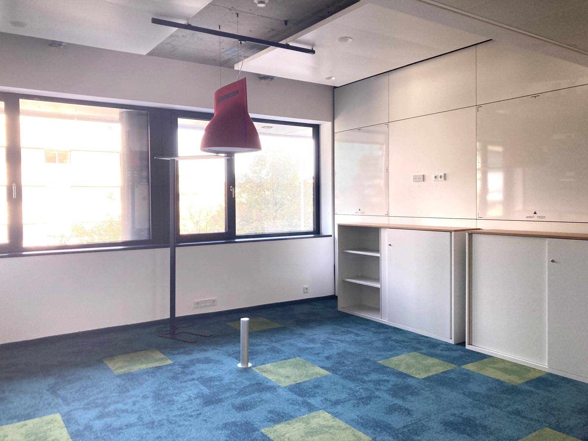 Helles Büro mit blau-grünem Teppichboden, Einbauschränken und großen Fenstern für viel Tageslicht.