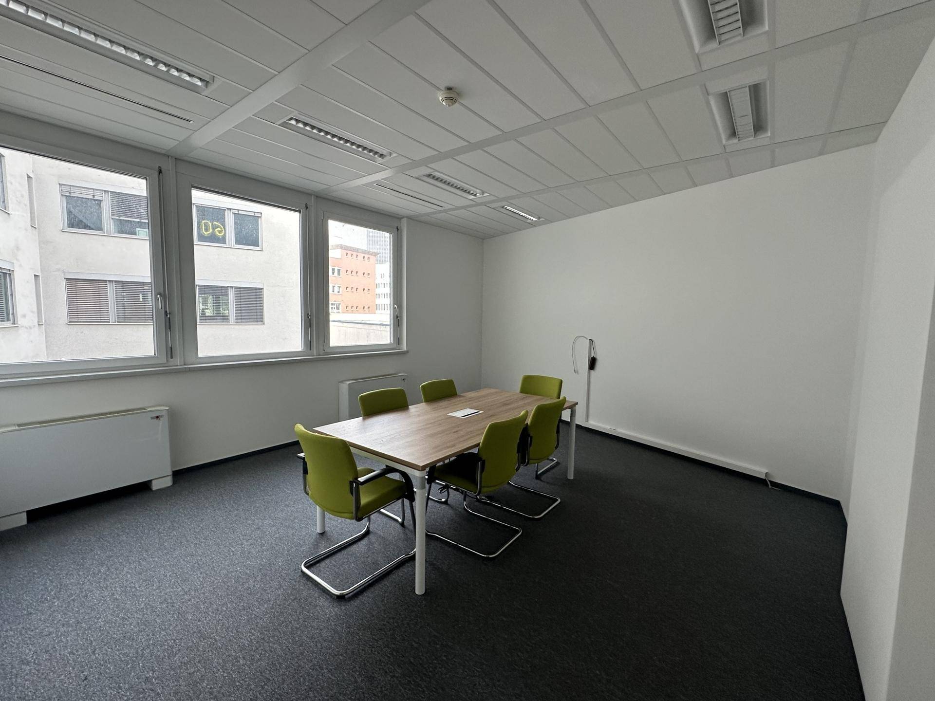 Konferenzraum mit großem Tisch, grünen Stühlen, Teppichboden und Fenstern mit Blick nach draußen.