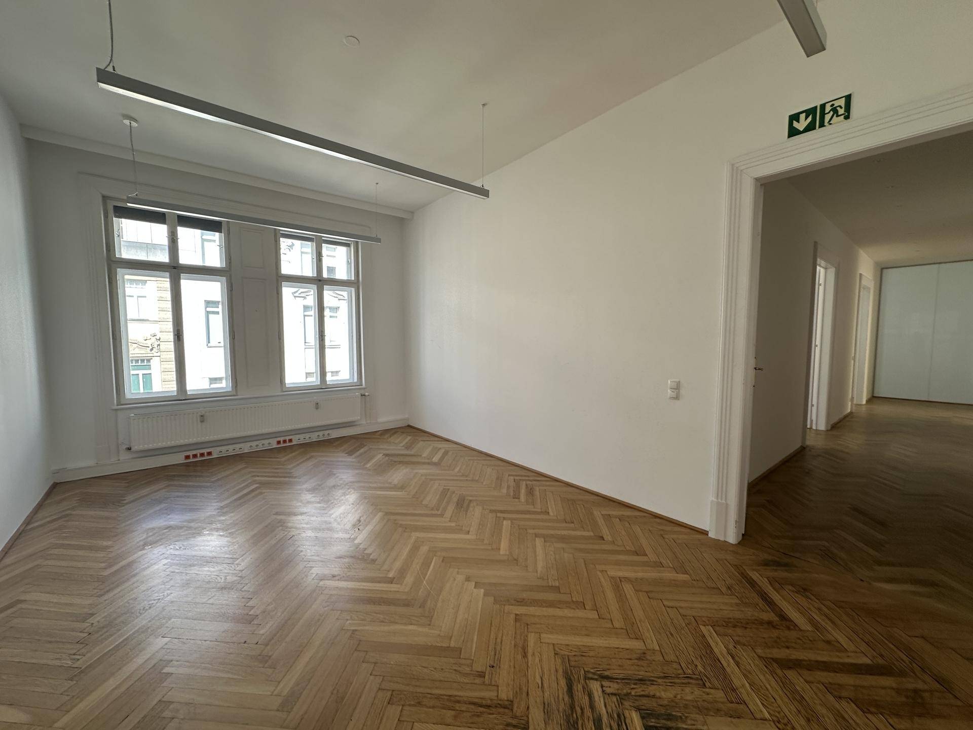 Heller, leerer Raum mit Fischgrätparkett und großen Fenstern, ideal für Büronutzung.