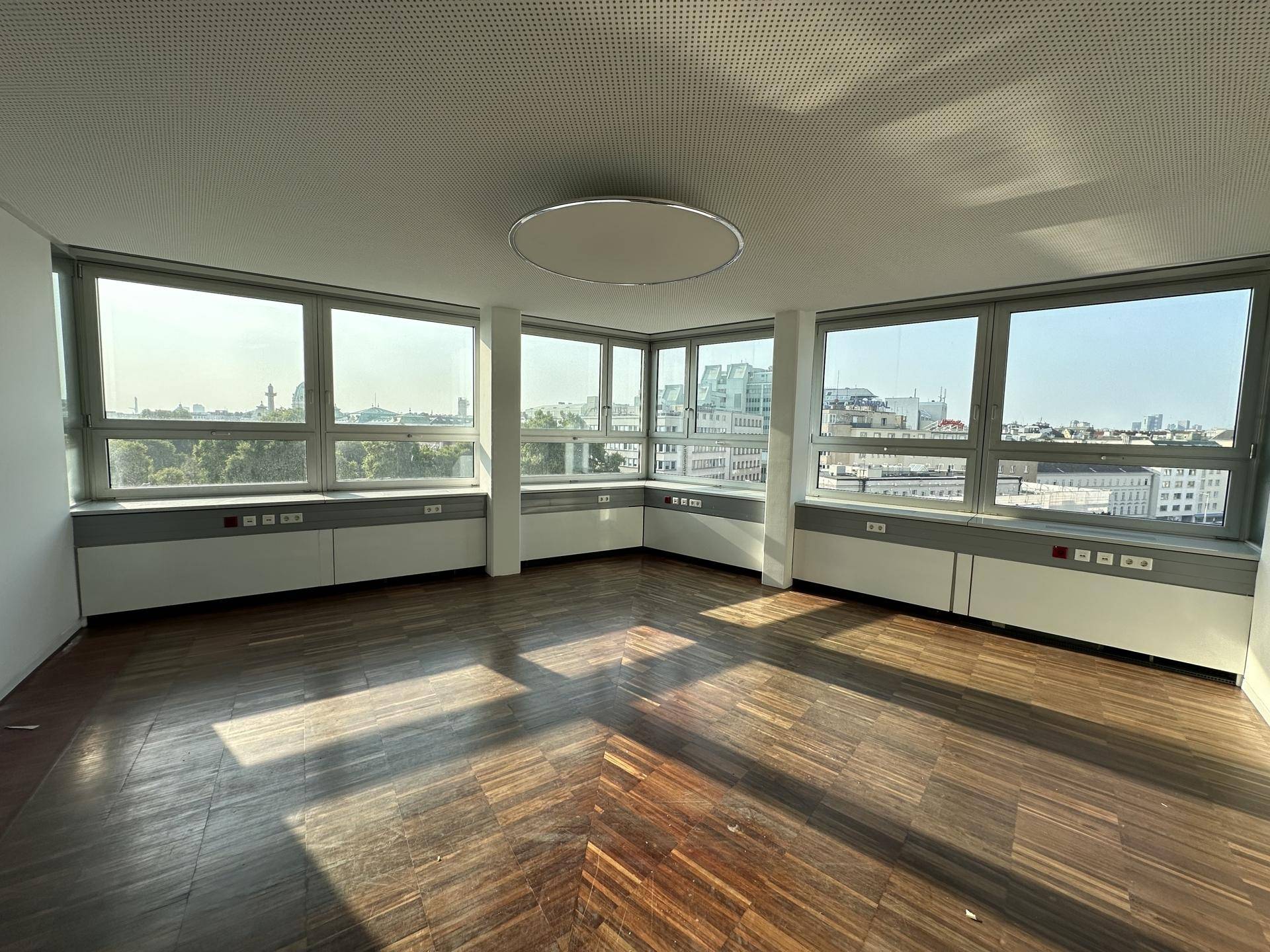 Heller Büroraum mit großen Fenstern, die viel Tageslicht hereinlassen, und einem dunklen Holzparkettboden.