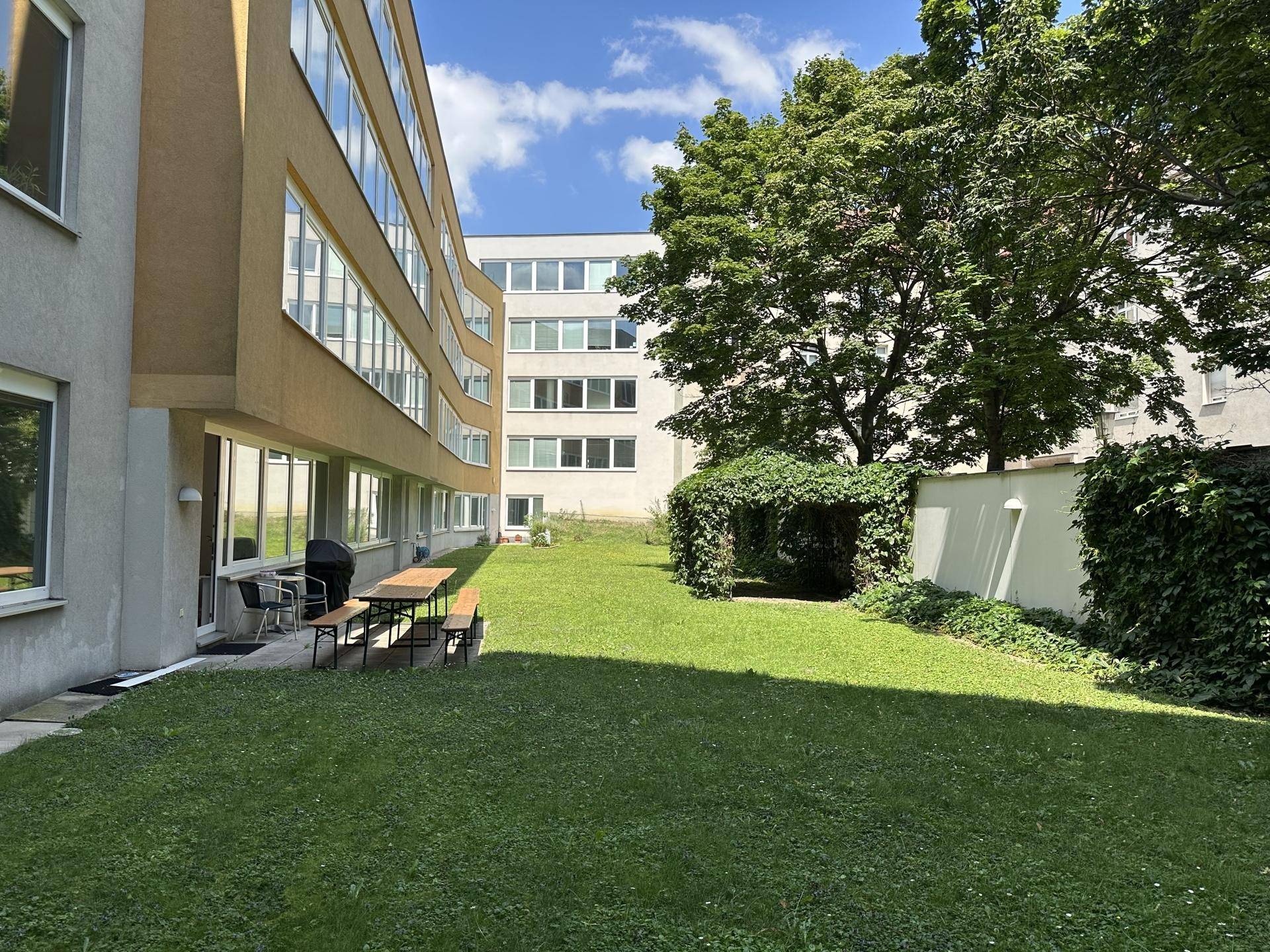 Grüner Gartenbereich mit Rasenfläche und Sitzgelegenheiten im Freien, umgeben von Gebäuden.