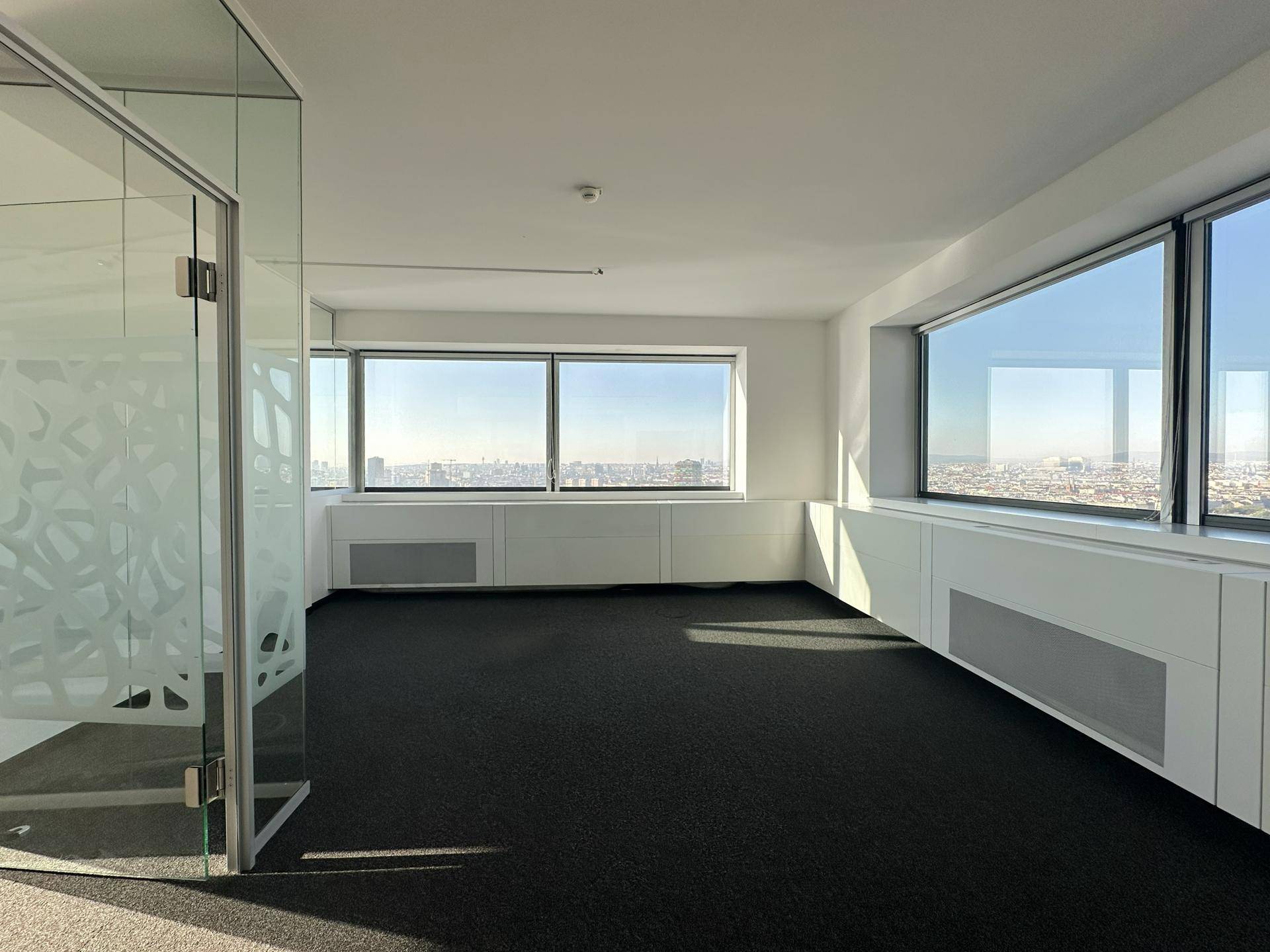 Geräumiges Büro mit dunklem Teppichboden, weißen Wänden und großen Fenstern mit Panoramablick auf die Stadt.
