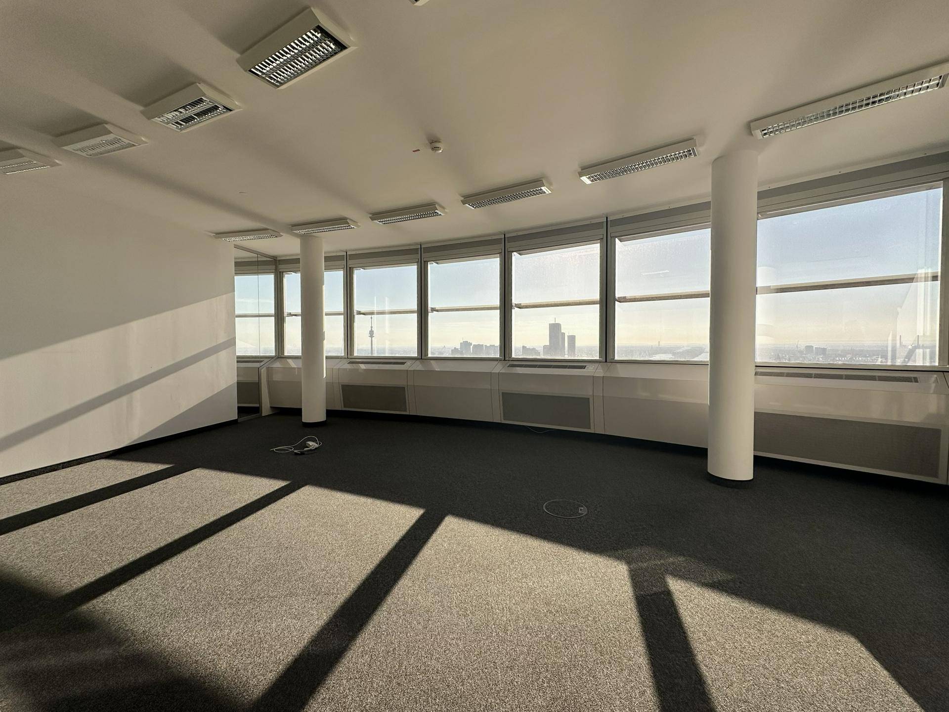 Leerer Büroraum mit grauem Teppichboden, weißen Wänden und einer Fensterfront mit Panoramablick auf die Stadt.