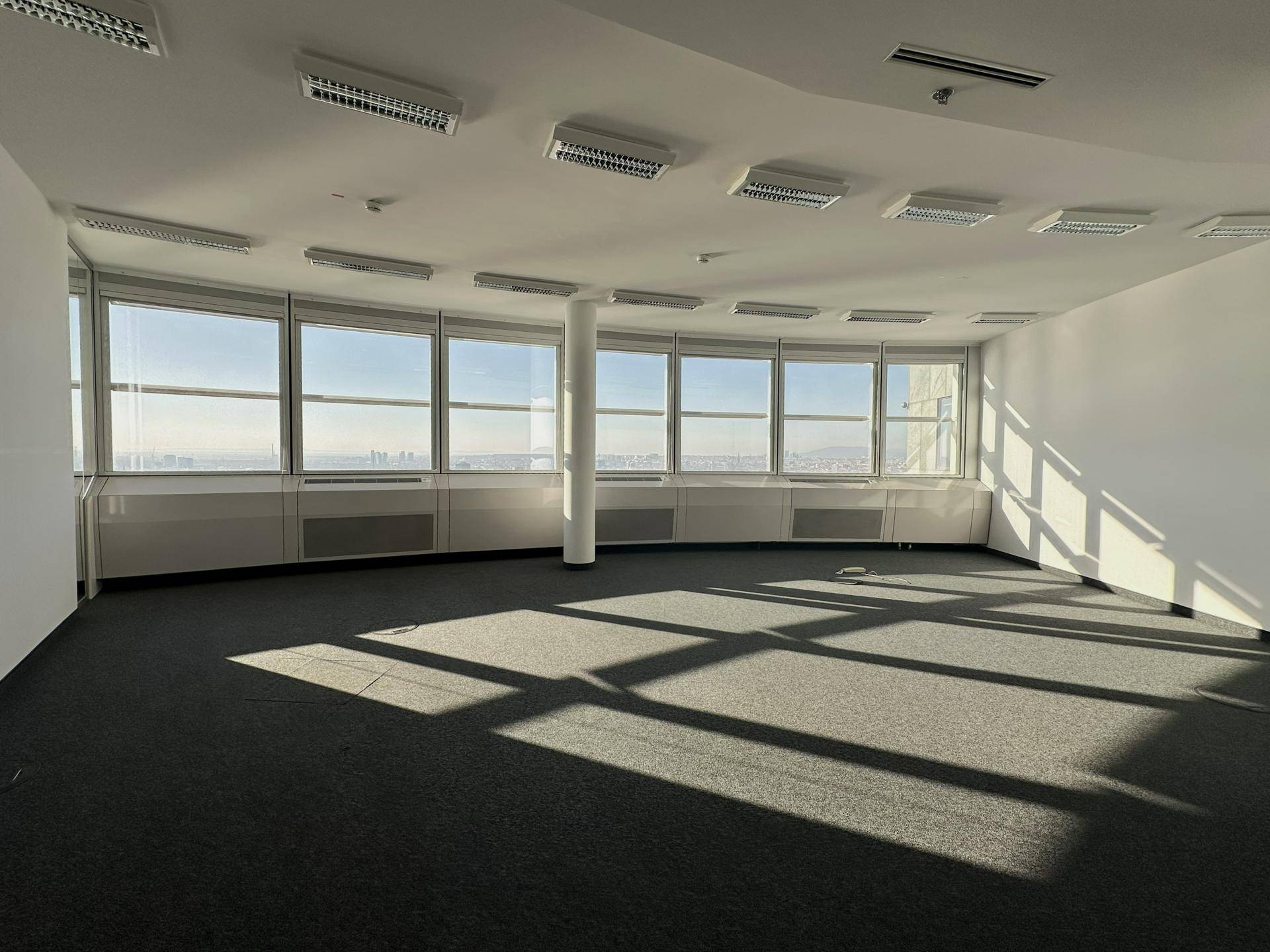 Geräumiger, leerer Bürobereich mit Teppichboden und einer geschwungenen Fensterfront, die viel Tageslicht hereinlässt.