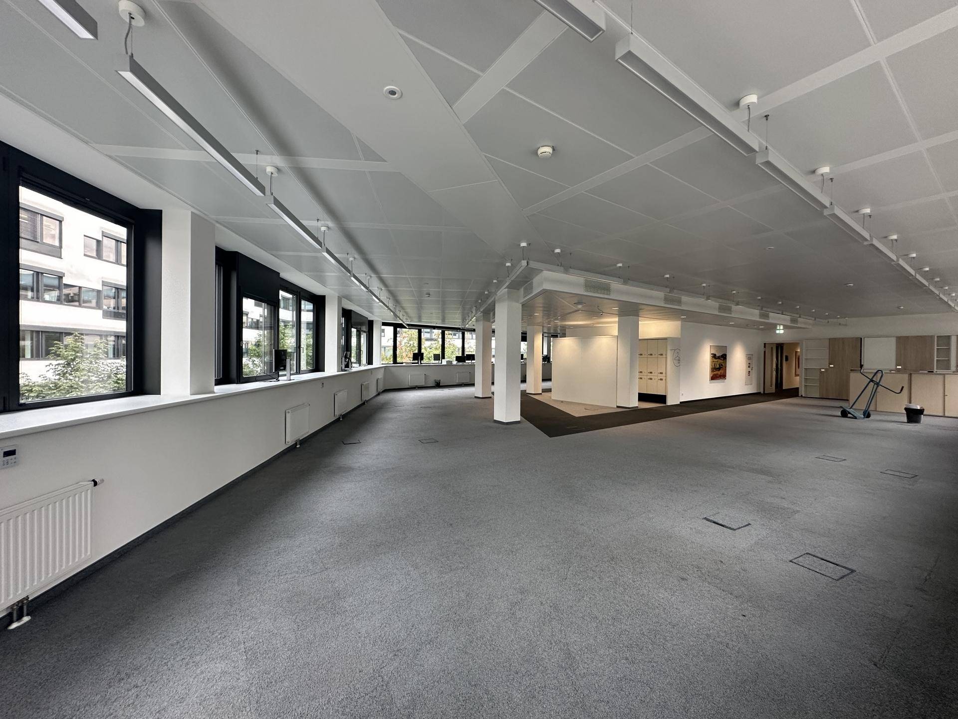 Großer, leerer Büroraum mit grauem Teppichboden, vielen Fenstern und moderner Deckenbeleuchtung.