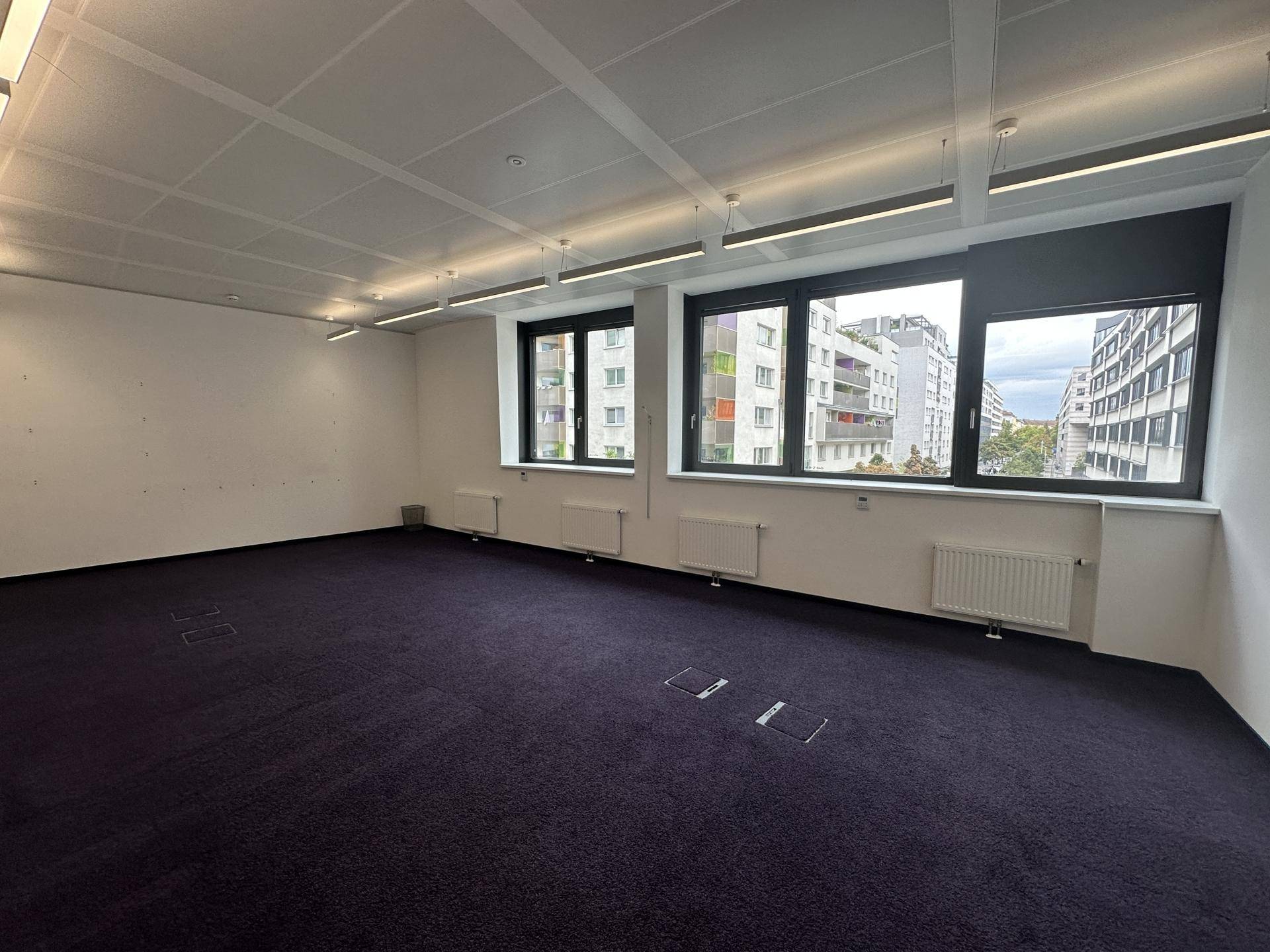 Leerer Büroraum mit dunklem Teppichboden und großen Fenstern, die einen Blick auf die Stadt bieten.