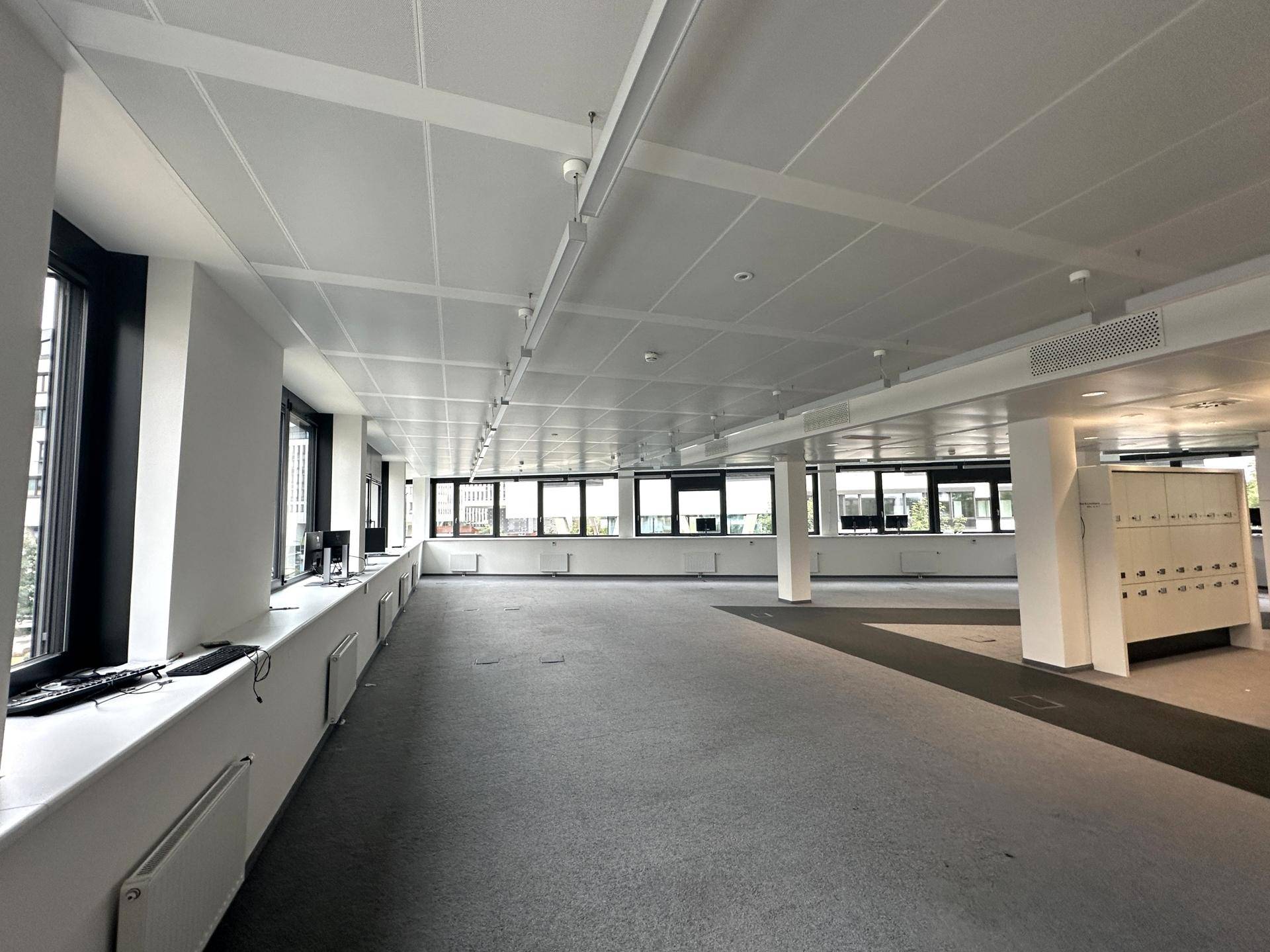 Heller Bürobereich mit grauen Teppichfliesen, langen Fensterfronten und Blick auf umliegende Gebäude.