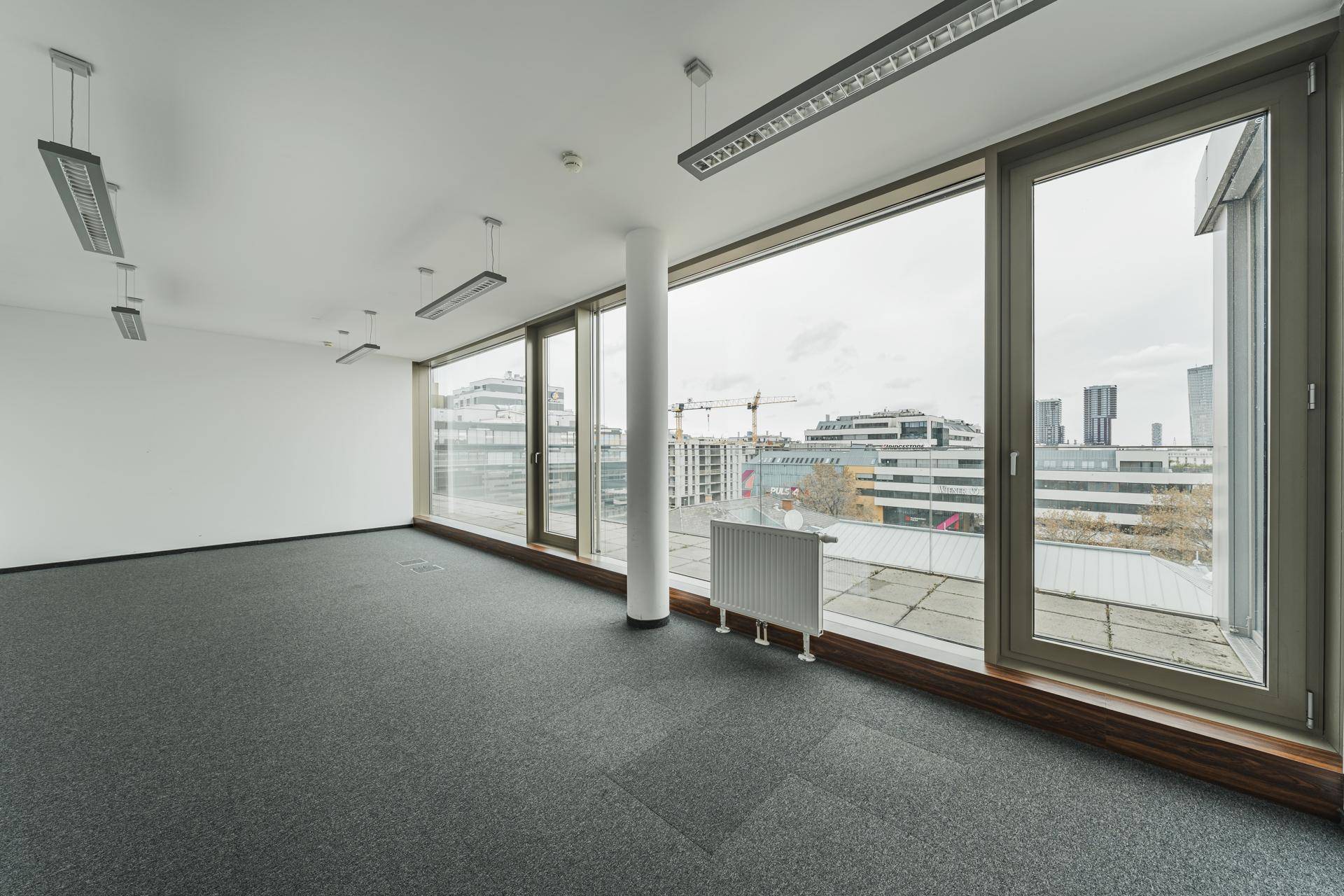 Leeres Büro mit Teppichboden, Säule und großen Fenstern mit Blick auf die Stadt.