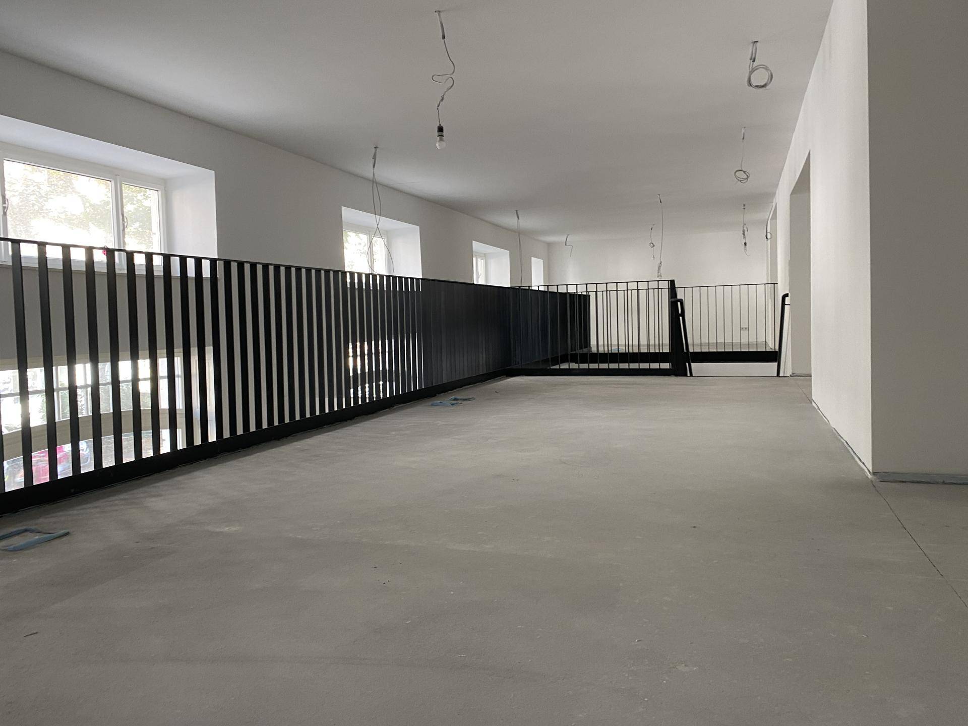 Großer, offener Innenraum mit Betonboden, weißen Wänden und schwarzem Metallgeländer, noch im Bau.