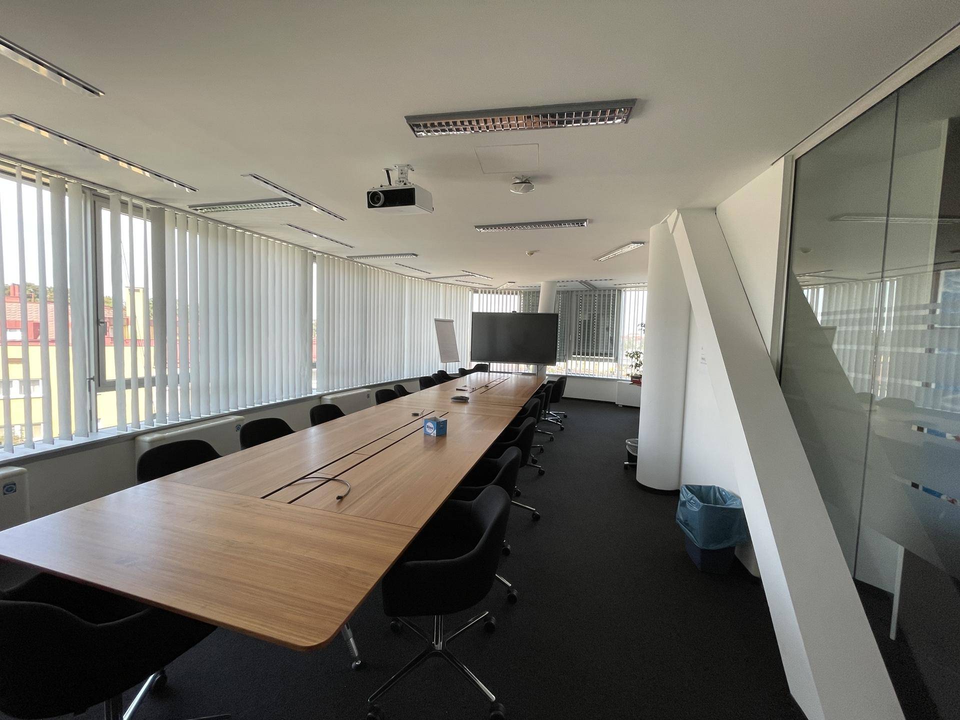 Geräumiger Konferenzraum mit langem Holztisch, vielen Stühlen, Beamer und großen Fenstern mit Jalousien.