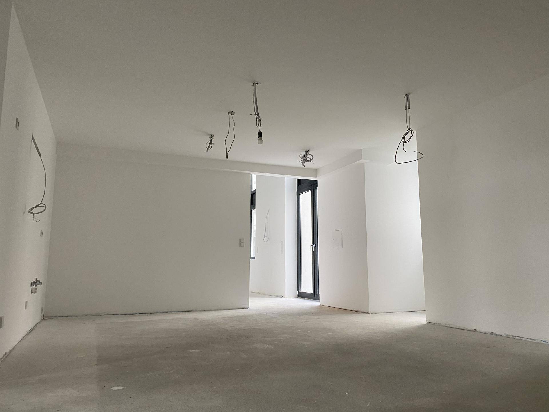 Offener Innenraum mit Betonboden, weißen Wänden und einer Tür oder einem großen Fenster, noch im Bau.