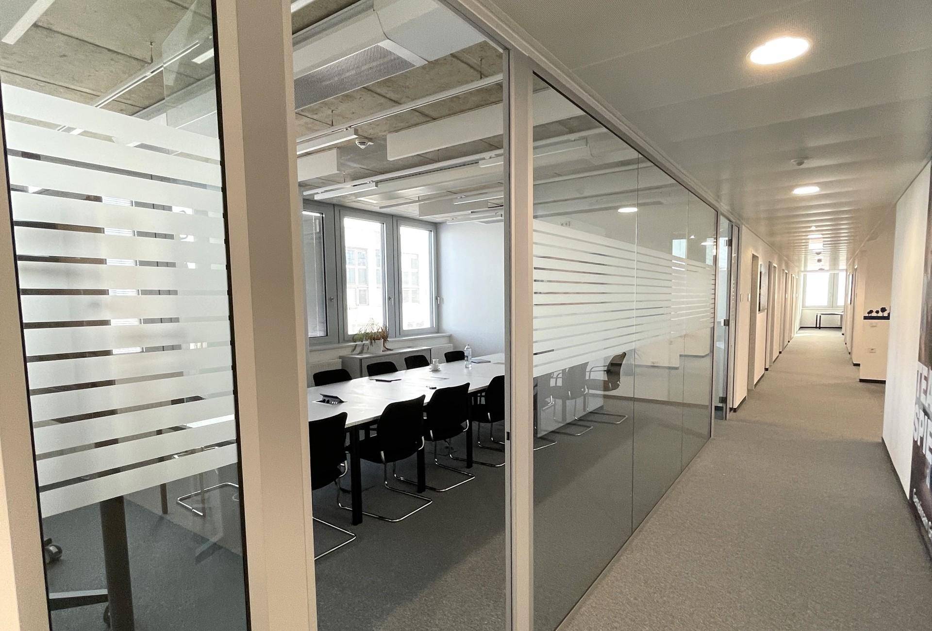 Heller Bürogang mit Glaswänden, die zu einem Konferenzraum führen, und Teppichboden.