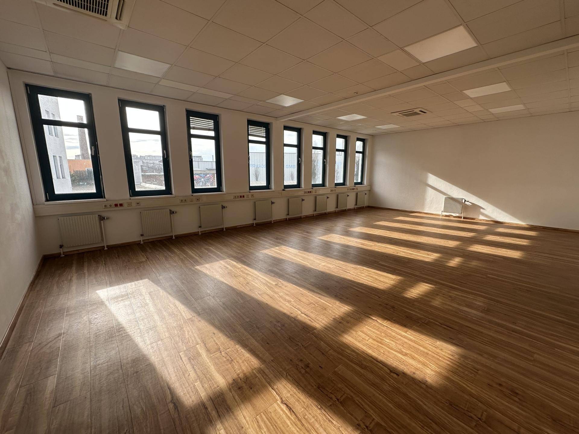 Heller Büroraum mit Holzboden und vielen Fenstern, durch die Sonnenlicht fällt.