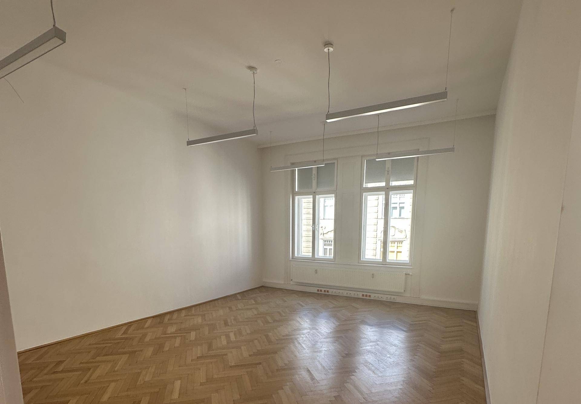 Heller, leerer Raum mit Fischgrätparkett, großen Fenstern und modernen Deckenleuchten.
