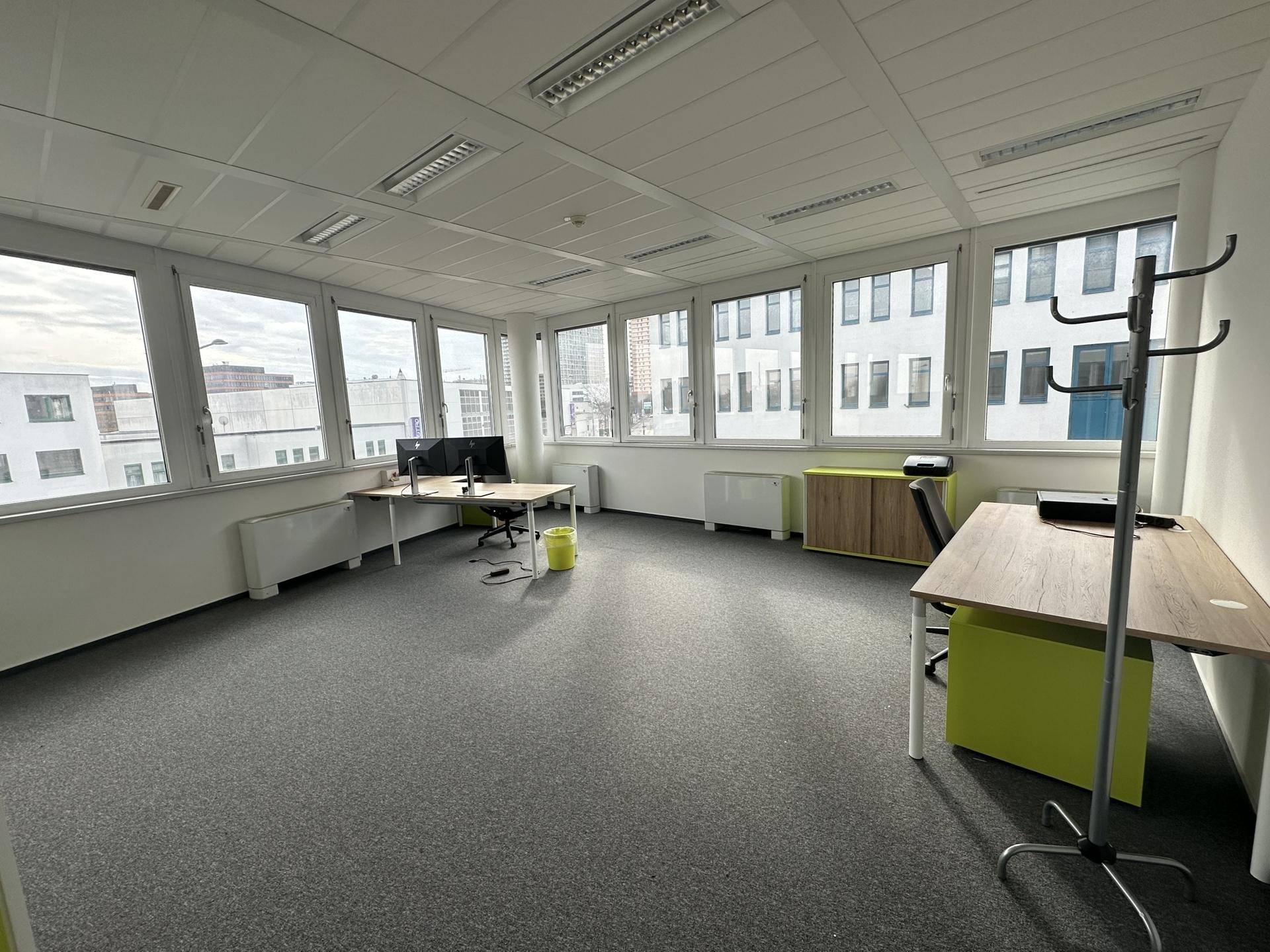 Helles Büro mit großen Fenstern, Teppichboden und modernen Schreibtischen mit Blick auf die städtische Umgebung.
