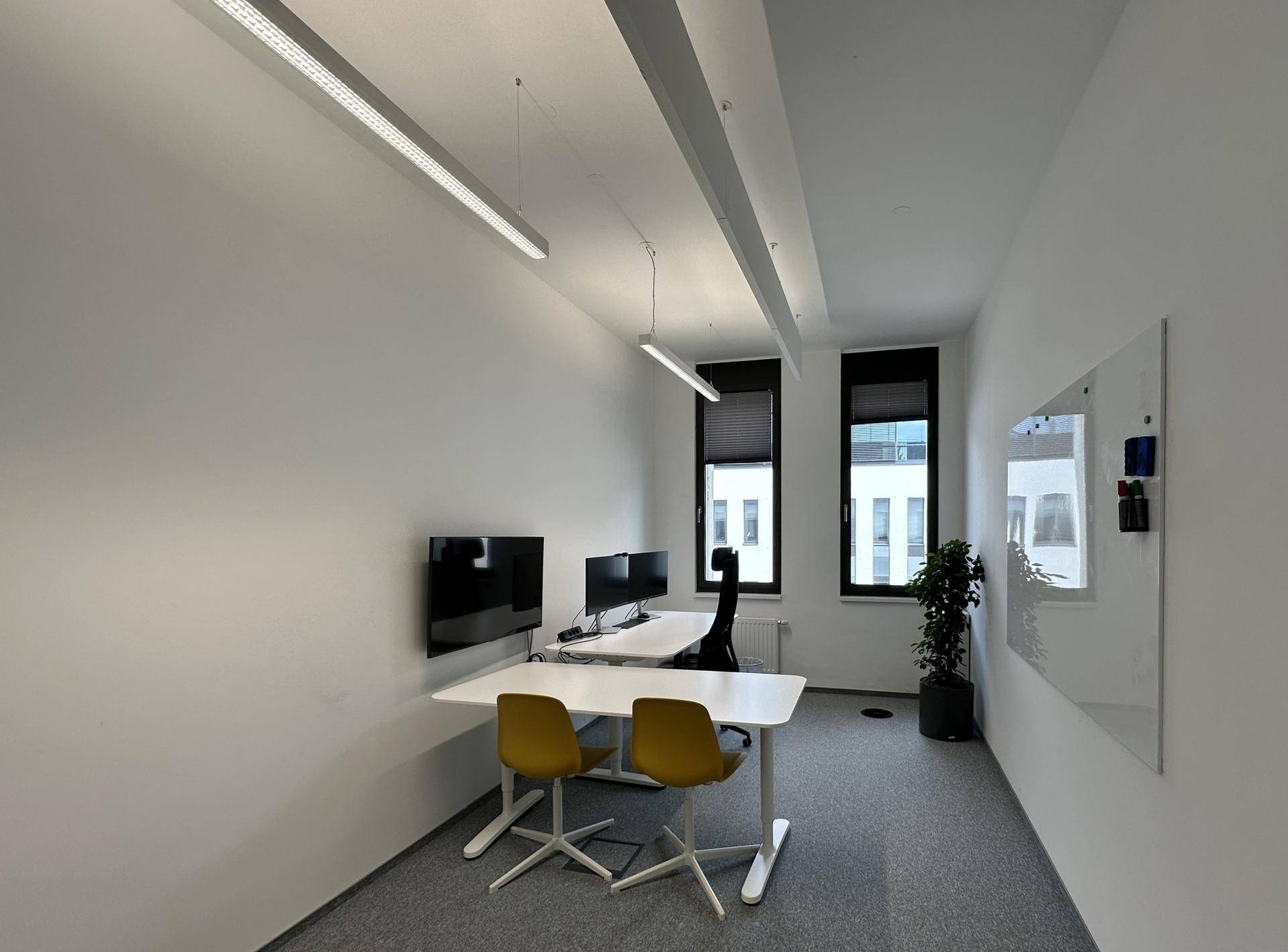 Modernes Büro mit zwei Arbeitsplätzen, gelben Stühlen, grauen Teppichböden und einer Zimmerpflanze am Fenster.