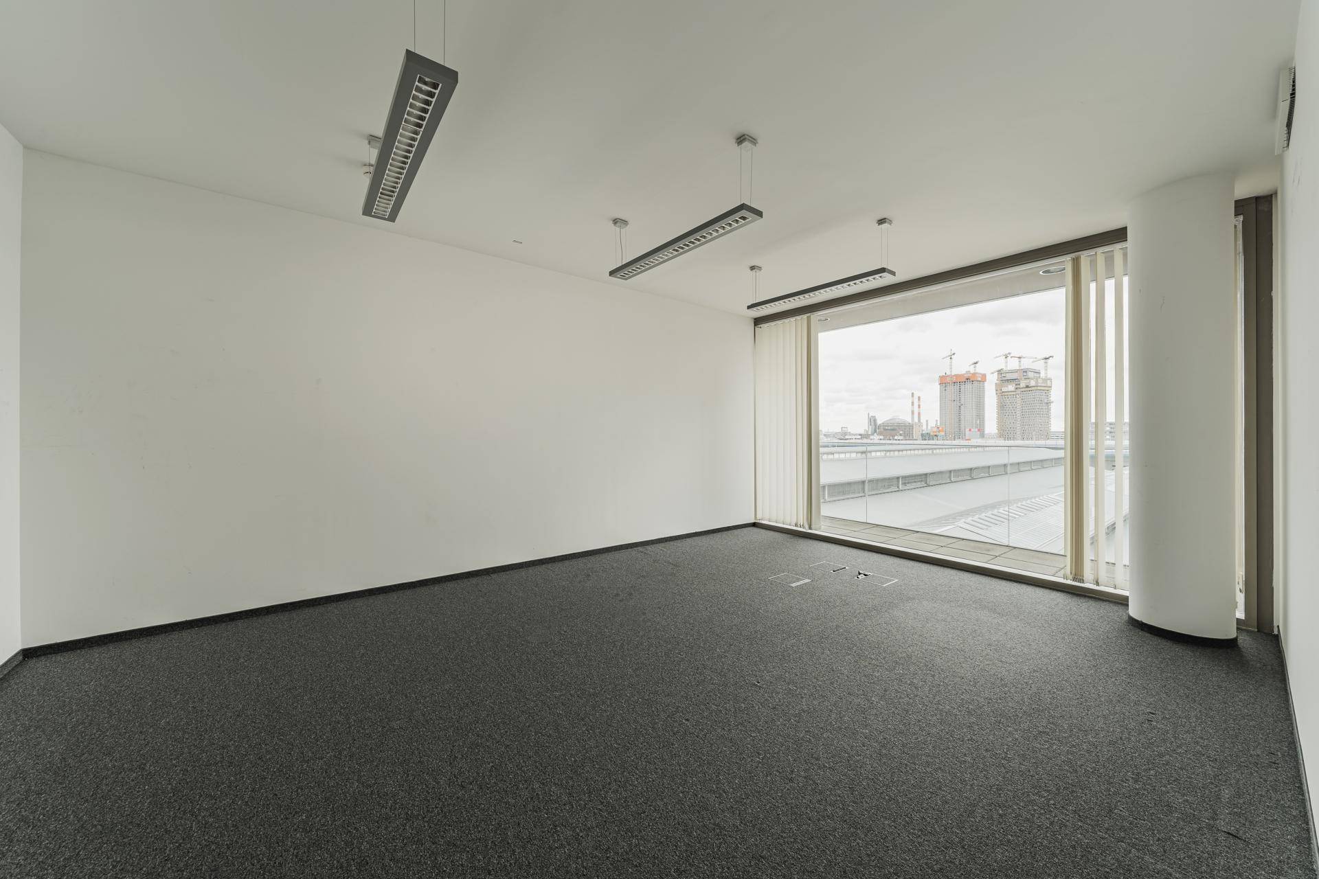 Helles Eckbüro mit Teppichboden und großen Fenstern, die viel Tageslicht hereinlassen.