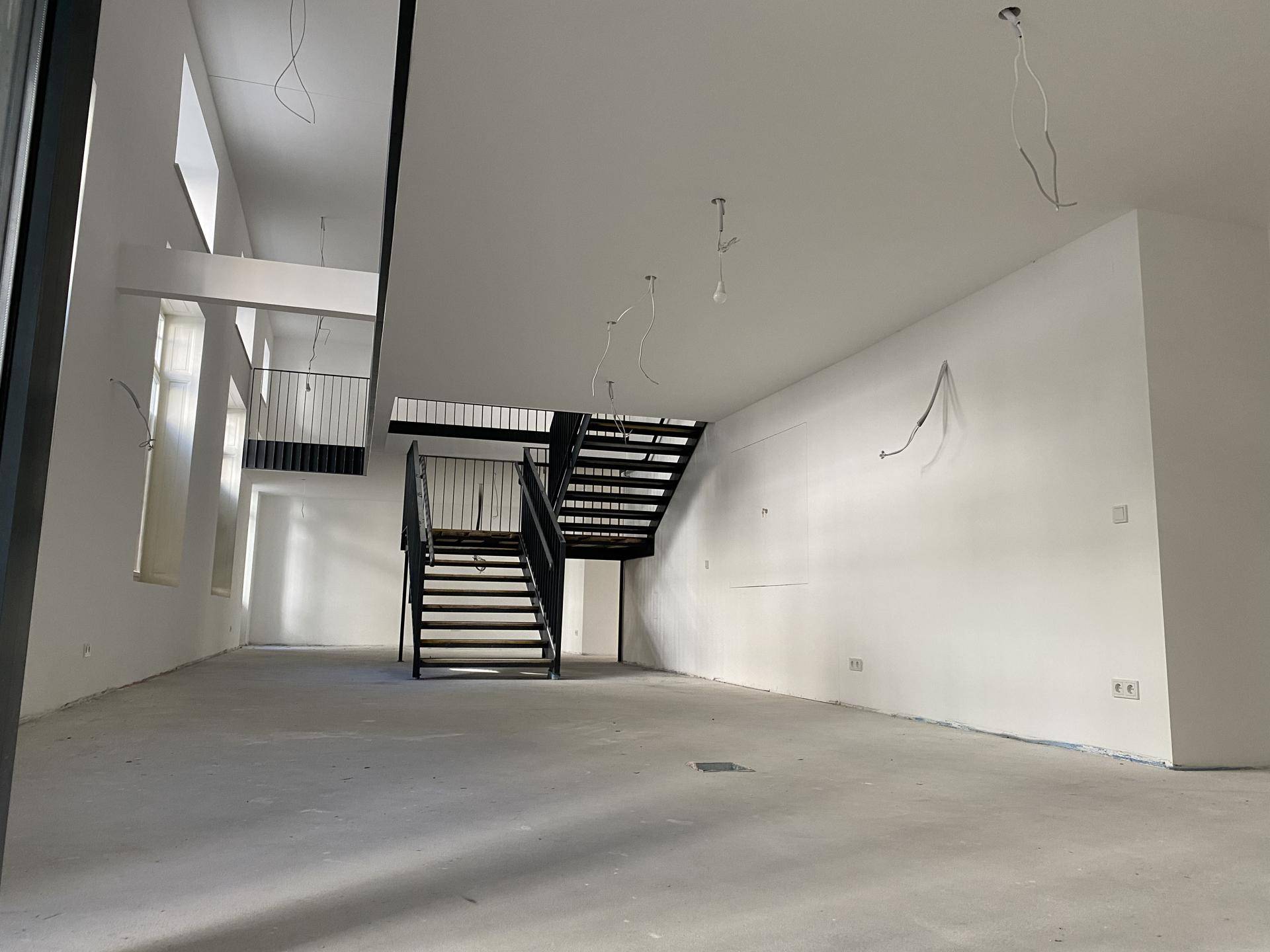 Geräumiger, offener Innenraum mit Betonboden, weiß gestrichenen Wänden und einer modernen schwarzen Metalltreppe.