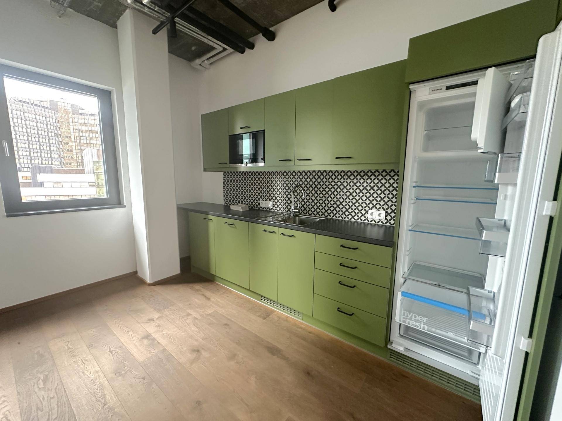 Moderne Küchenzeile in Grün mit Kühlschrank, Mikrowelle und Holzboden in einem Büroraum.