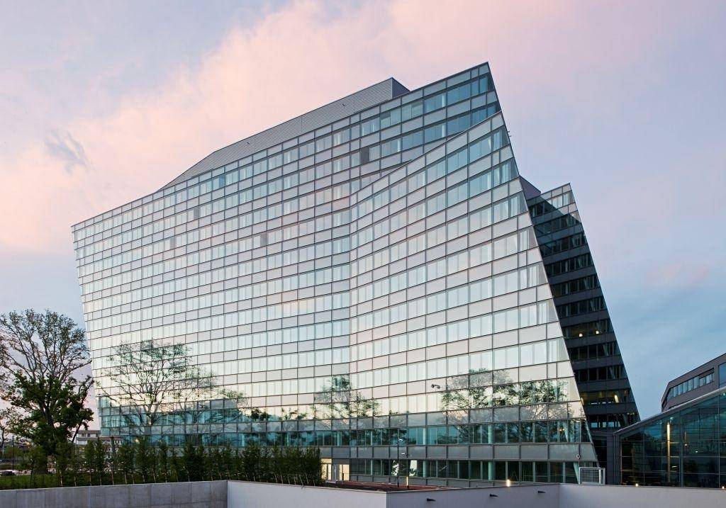 Großes, modernes Gebäude mit wellenförmiger Glasfassade, das den Himmel und die Bäume widerspiegelt.