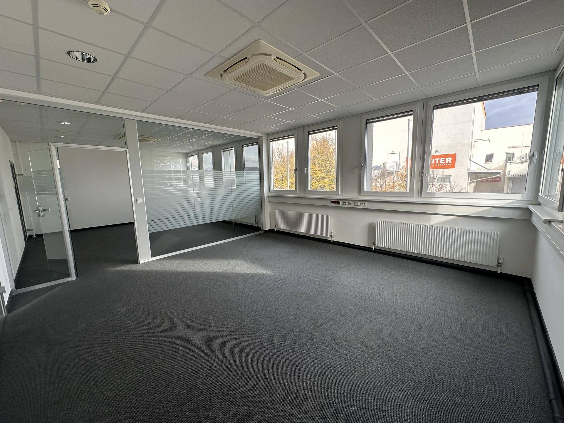 Geräumiges Büro mit Teppichboden, großen Fenstern und einer Glaswand zu einem weiteren Raum.