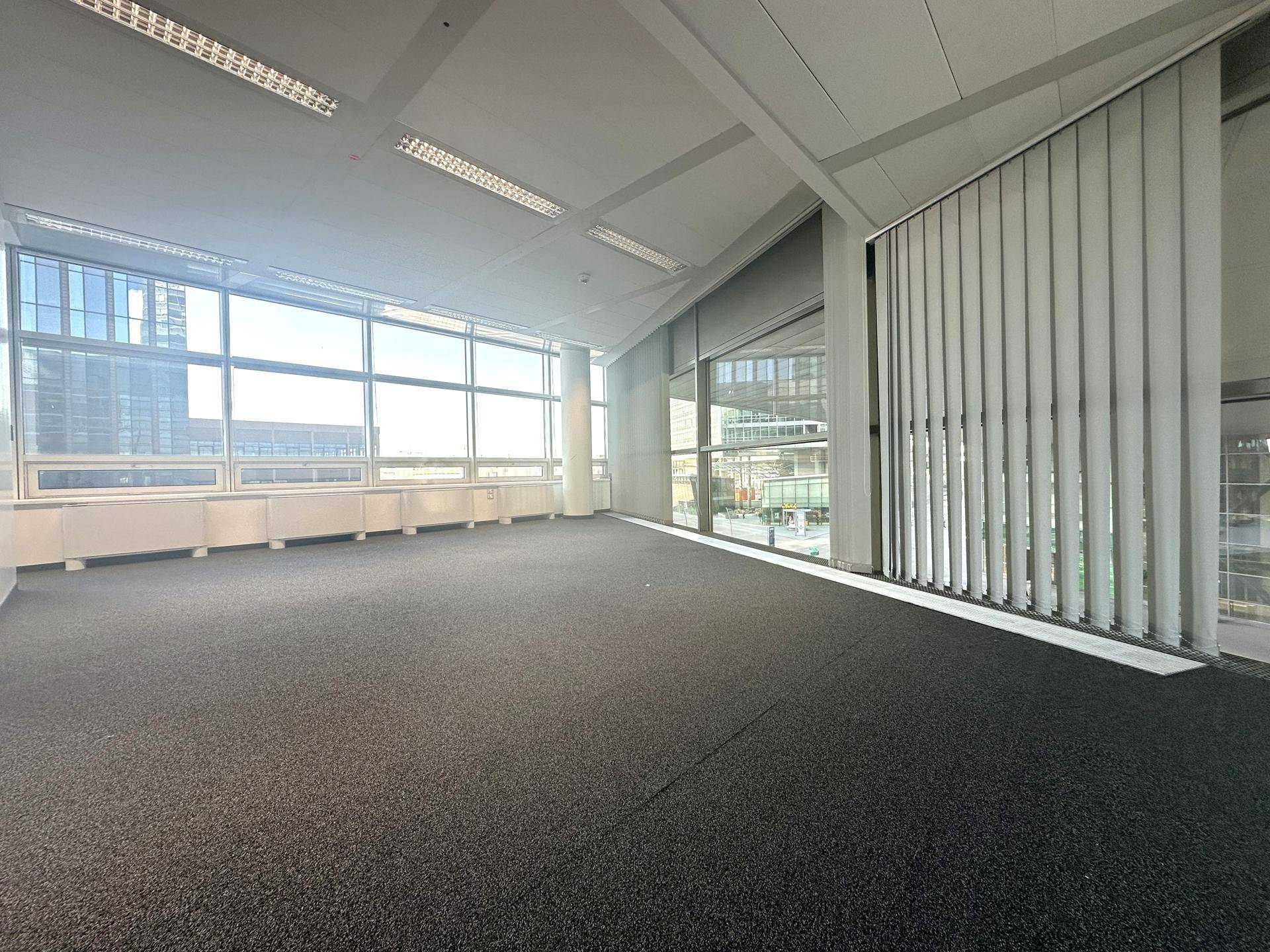 Helles Büro mit Teppichboden und vertikalen Jalousien an den großen Fenstern.