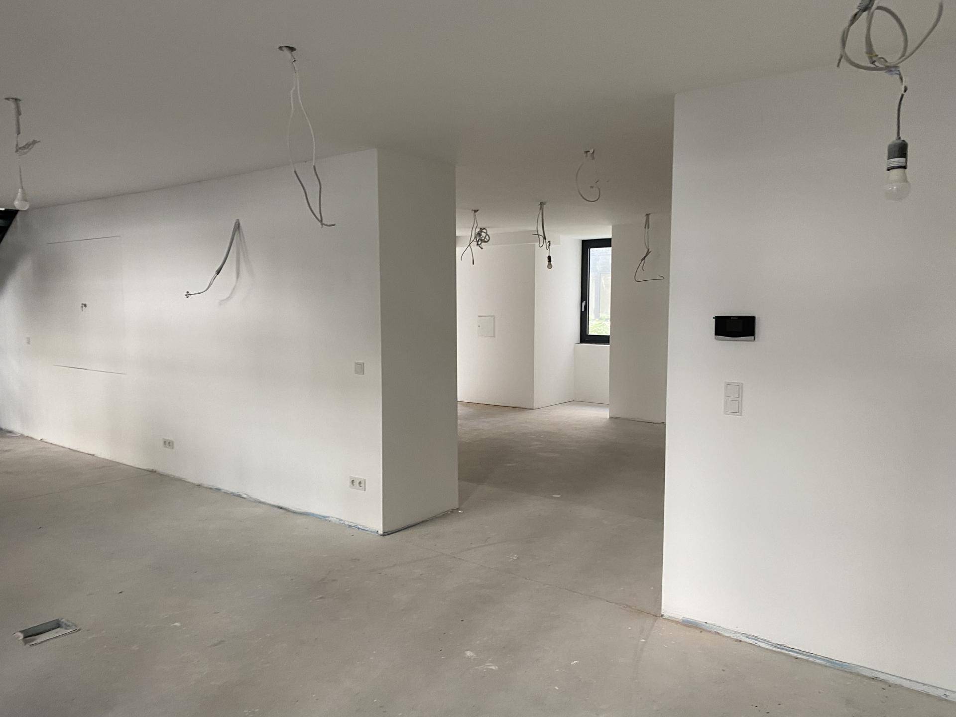 Heller, offener Innenraum mit Betonboden, weißen Wänden und einem einzelnen Fenster, noch im Rohbau.