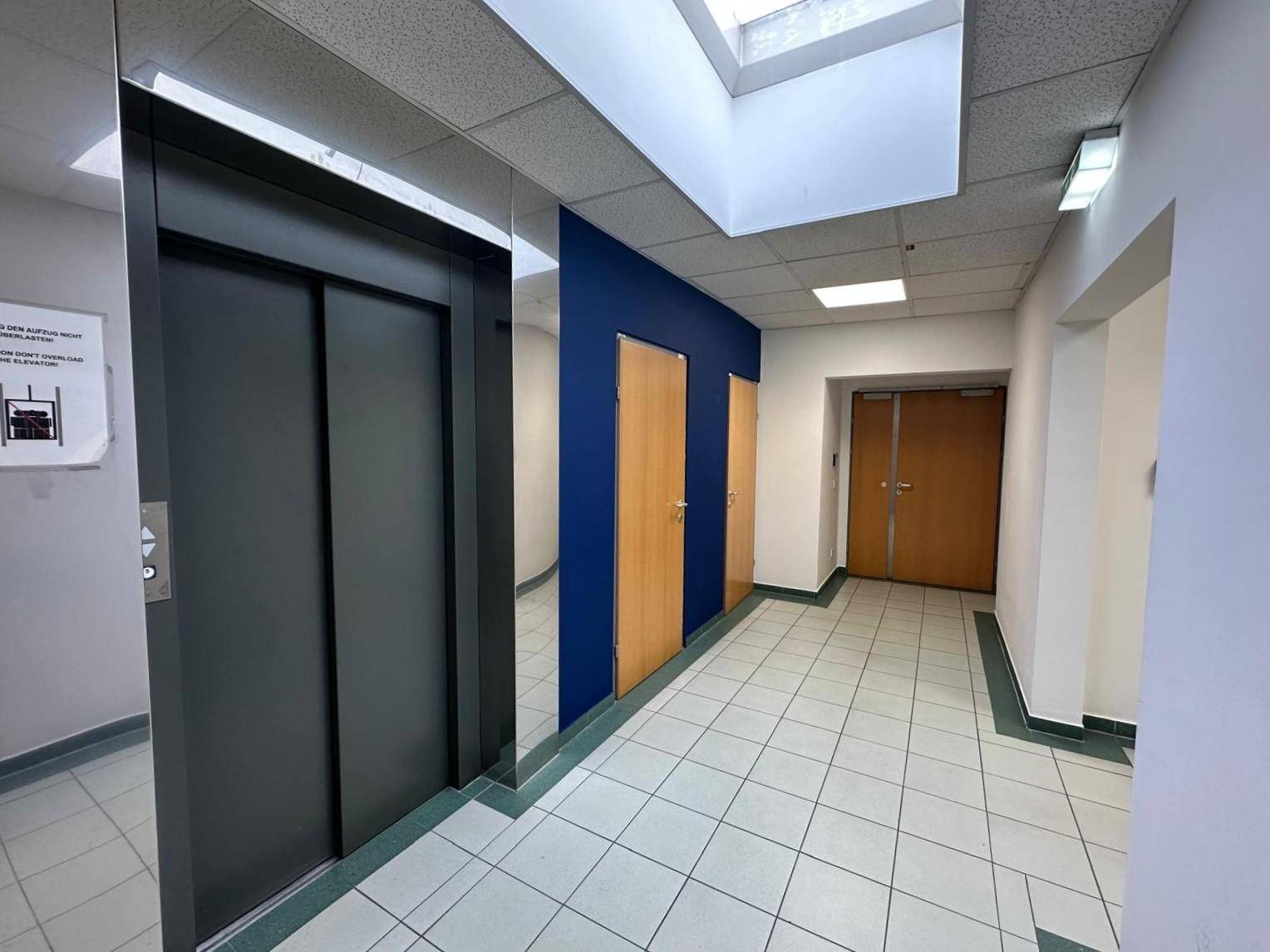 Heller Flur mit Aufzug, blauer Akzentwand und Fliesenboden in einem Bürogebäude.