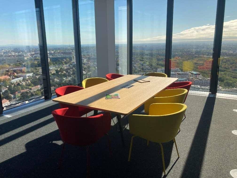Heller Konferenzraum mit modernem Holztisch und farbigen Stühlen, umgeben von großen Fenstern mit Stadtpanorama.