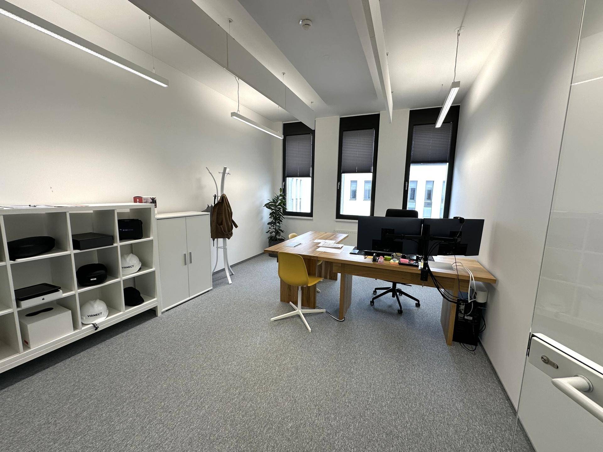 Geräumiges Büro mit Holzschreibtisch, gelbem Stuhl, weißen Regalen und grauen Teppichböden, gut beleuchtet.