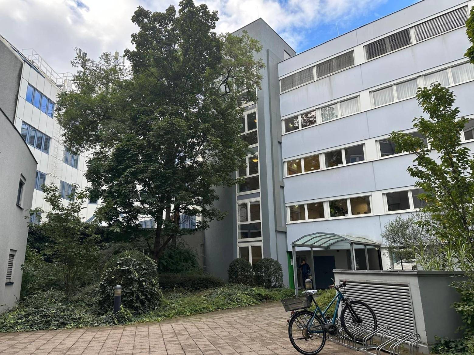 Eingangsbereich eines Bürogebäudes mit viel Grün, einem Baum und einem Fahrradständer im Hof.