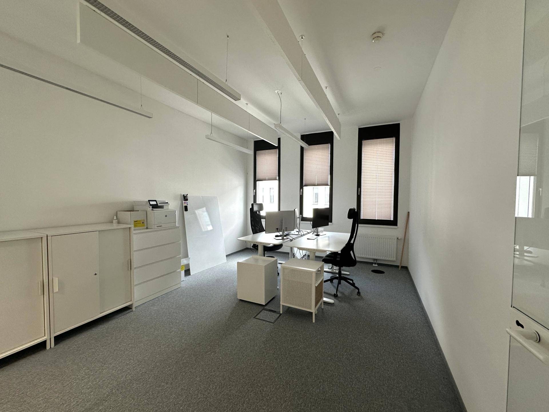 Helles, modernes Büro mit grauen Teppichböden, weißen Wänden und zwei Arbeitsplätzen mit Blick nach draußen.