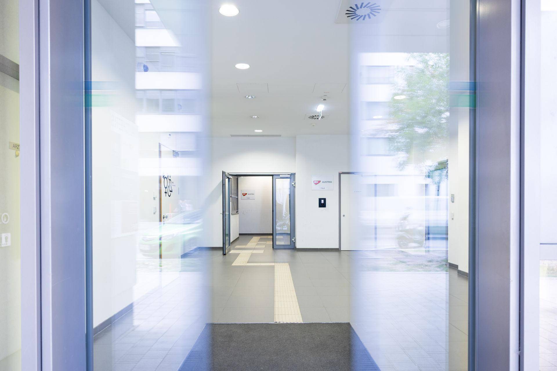 Heller Eingangsbereich mit großen Glastüren, weißen Wänden und grauem Fliesenboden, der nach draußen führt.