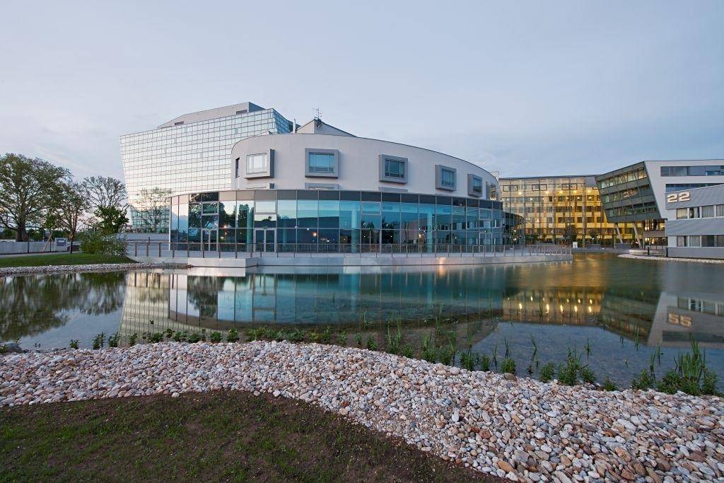 Modernes, geschwungenes Gebäude am Wasser, dessen Fassade sich im Teich spiegelt, umgeben von Grünflächen.