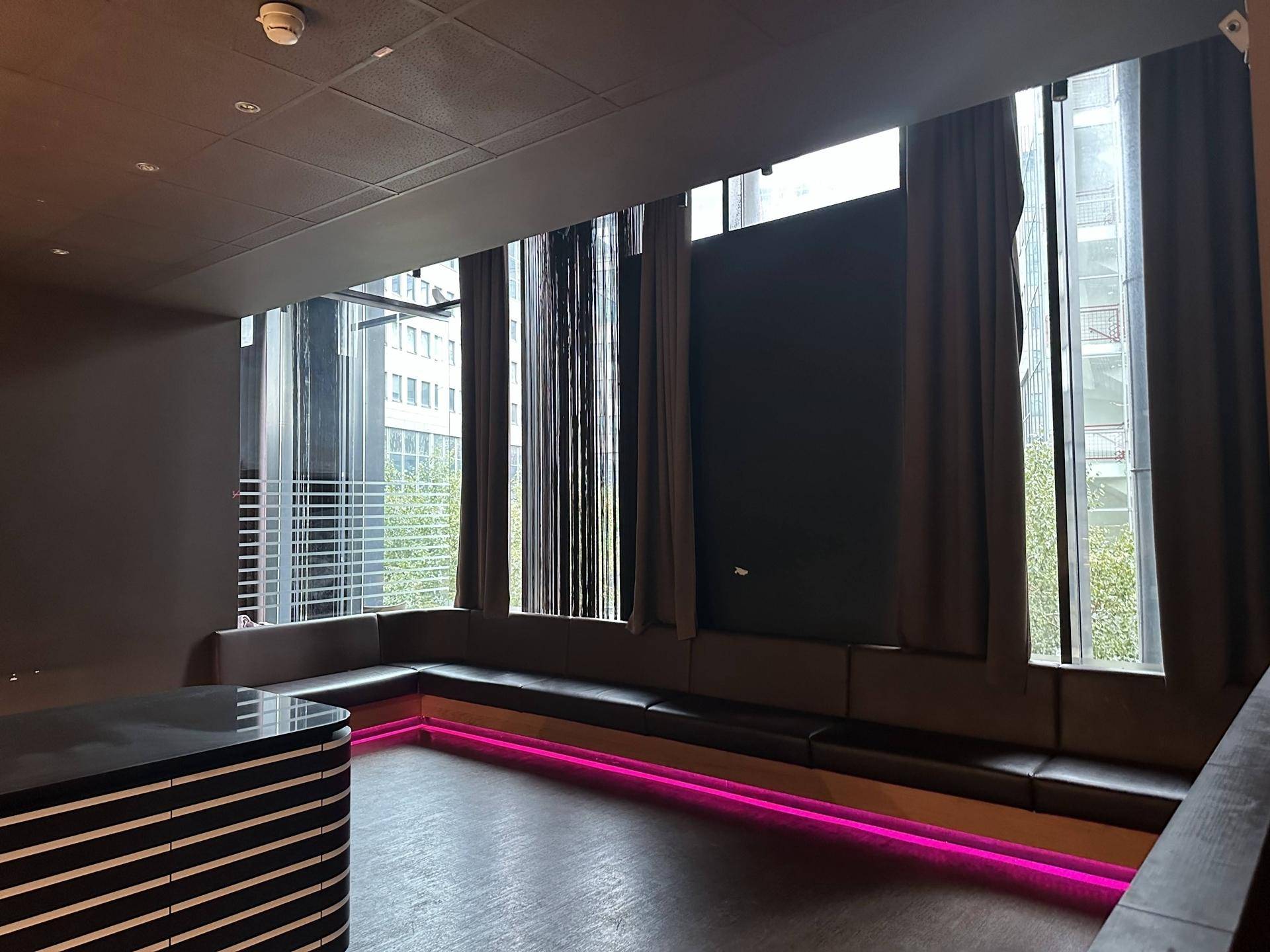 Gemütlicher Loungebereich mit langer Sitzbank, großen Fenstern mit Vorhängen und moderner pinker LED-Beleuchtung.