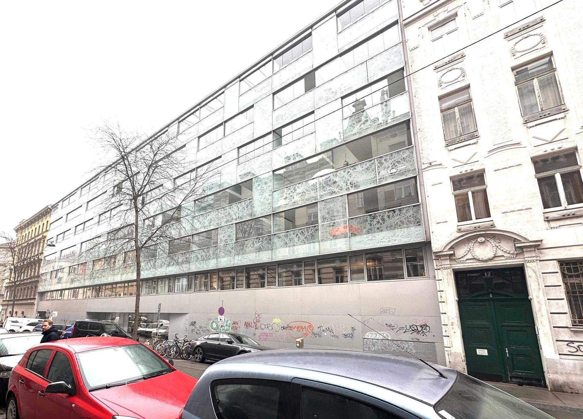 Modernes Bürogebäude mit großer Glasfassade an einer belebten Straße, umgeben von anderen Gebäuden.