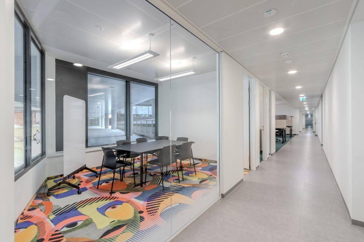 Heller Büroflur mit Glaswänden, der zu einem modernen Besprechungsraum mit farbenfrohem Teppich führt.