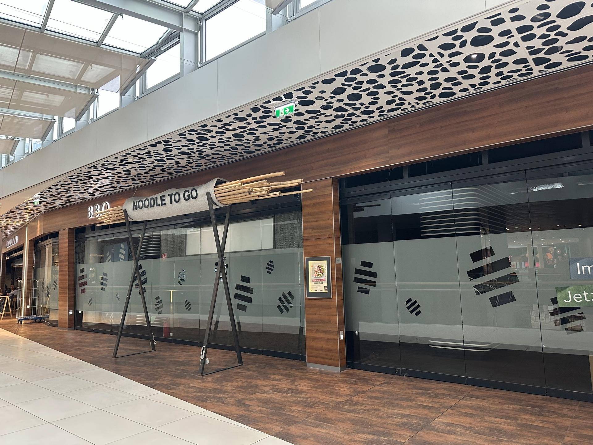 Außenansicht eines "Noodle to Go"-Restaurants in einem Einkaufszentrum mit großen Glasfronten und Holzverkleidung.