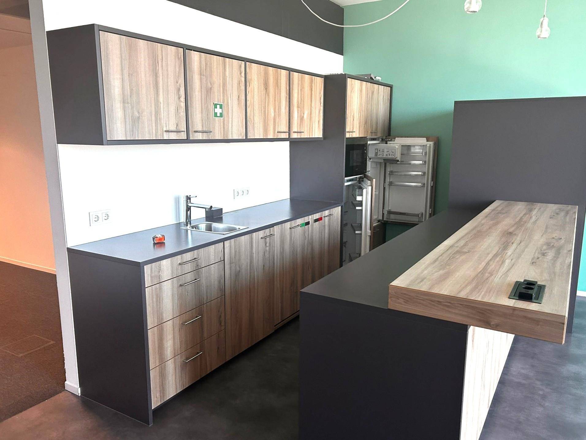 Moderne Teeküche mit Holzschränken, dunklen Arbeitsplatten und integriertem Kühlschrank in einem Bürobereich.