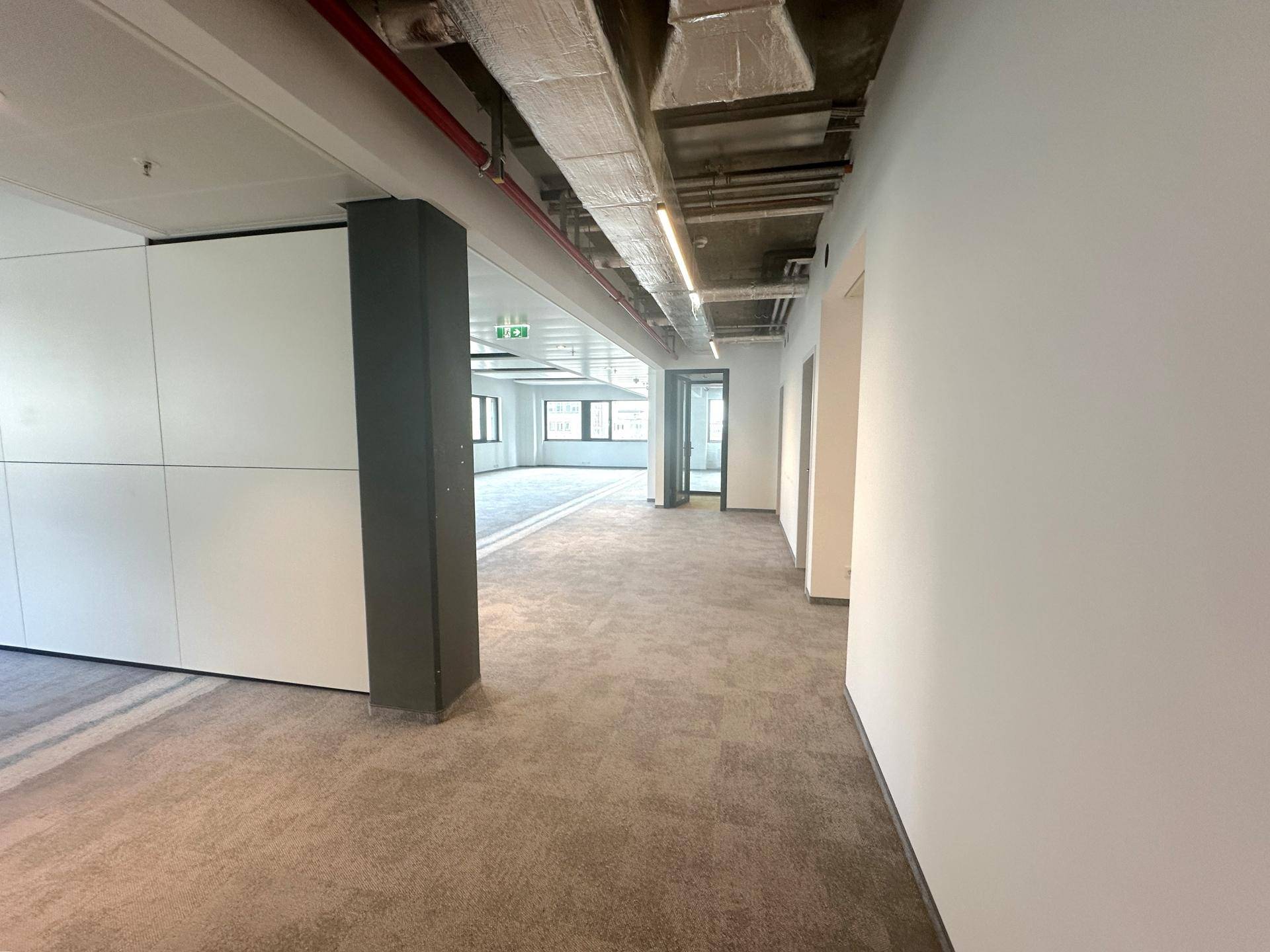 Langer Flur in einem modernen Bürogebäude mit Teppichboden und sichtbaren Lüftungsrohren.