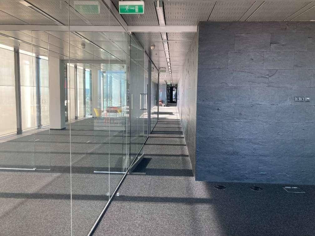 Moderner Büroflur mit Glaswänden auf einer Seite und einer dunklen Steinwand auf der anderen, mit Teppichboden.