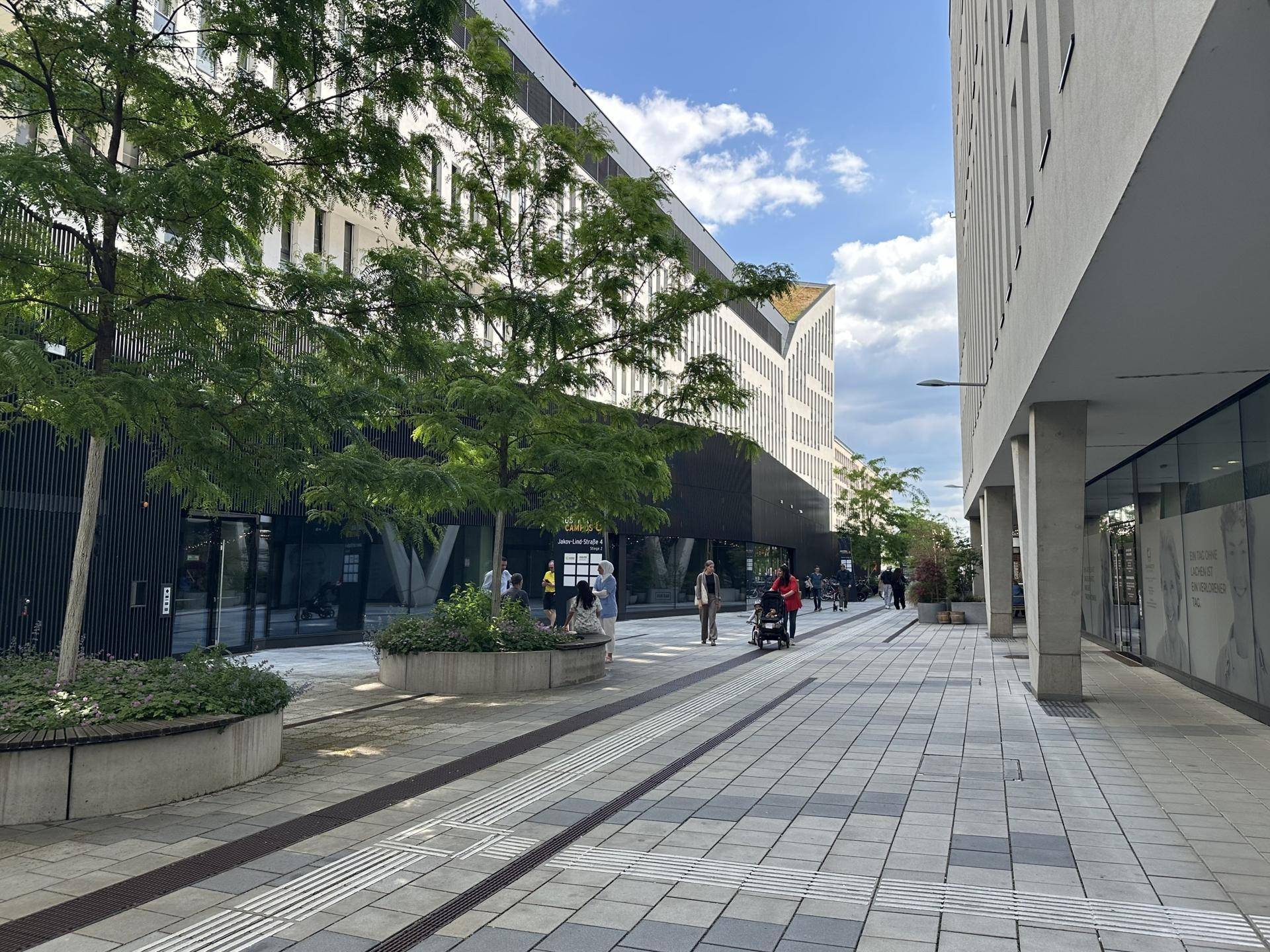 Belebte moderne Straße mit Bäumen, Sitzgelegenheiten und zeitgenössischen Gebäuden unter blauem Himmel.