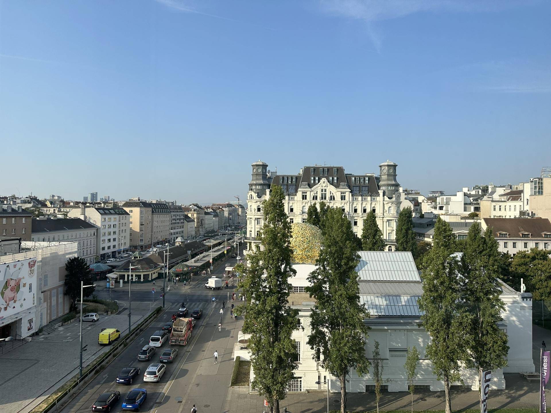 Weitläufige Stadtansicht von oben mit historischen Gebäuden, Bäumen und einer belebten Straße unter blauem Himmel.