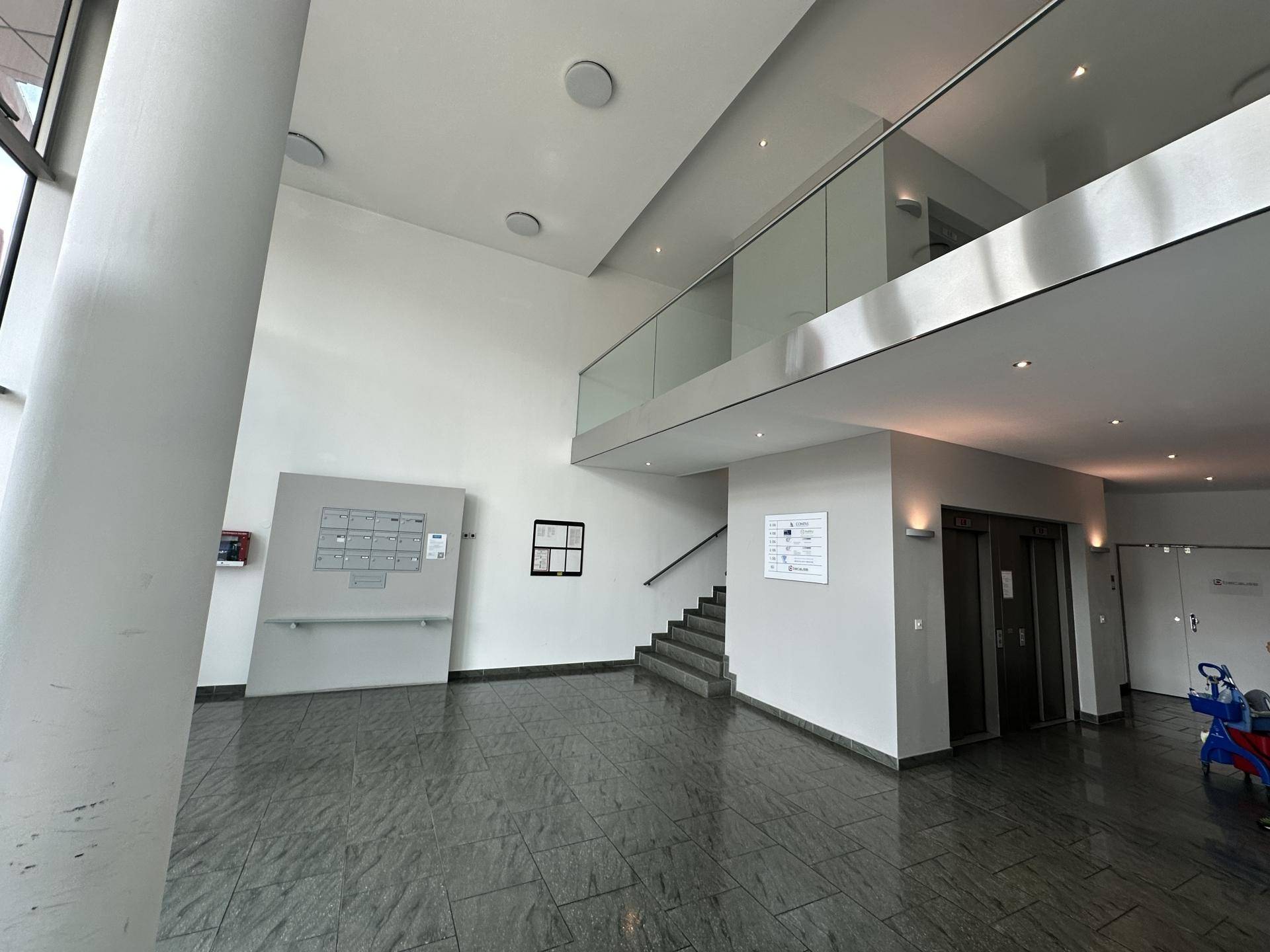 Geräumige, moderne Lobby mit dunklem Fliesenboden, hohen Decken und Glasgeländer zur oberen Etage.