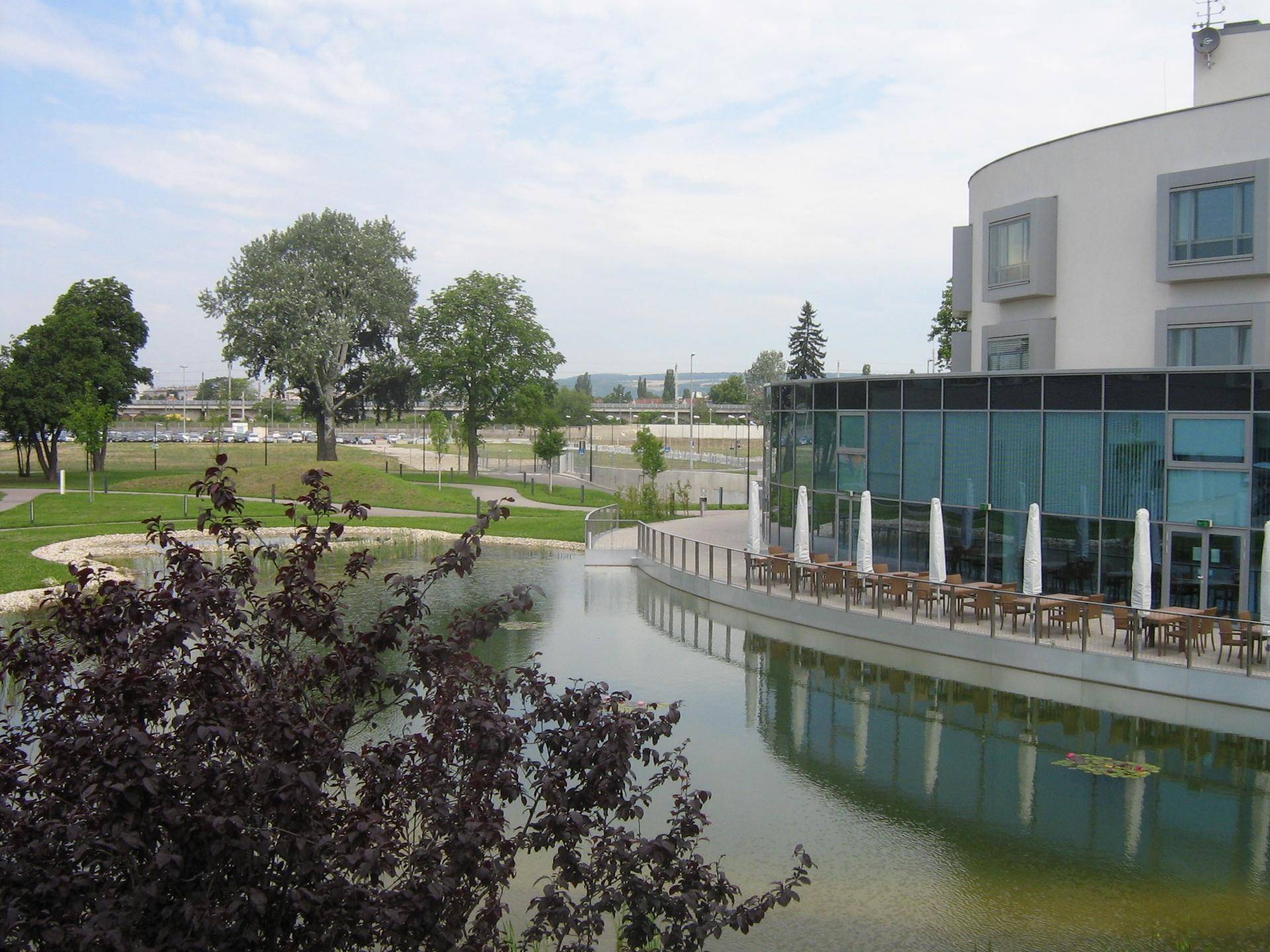 Modernes Gebäude mit geschwungener Glasfassade und einer Terrasse am Wasser, umgeben von Grünflächen und Bäumen.
