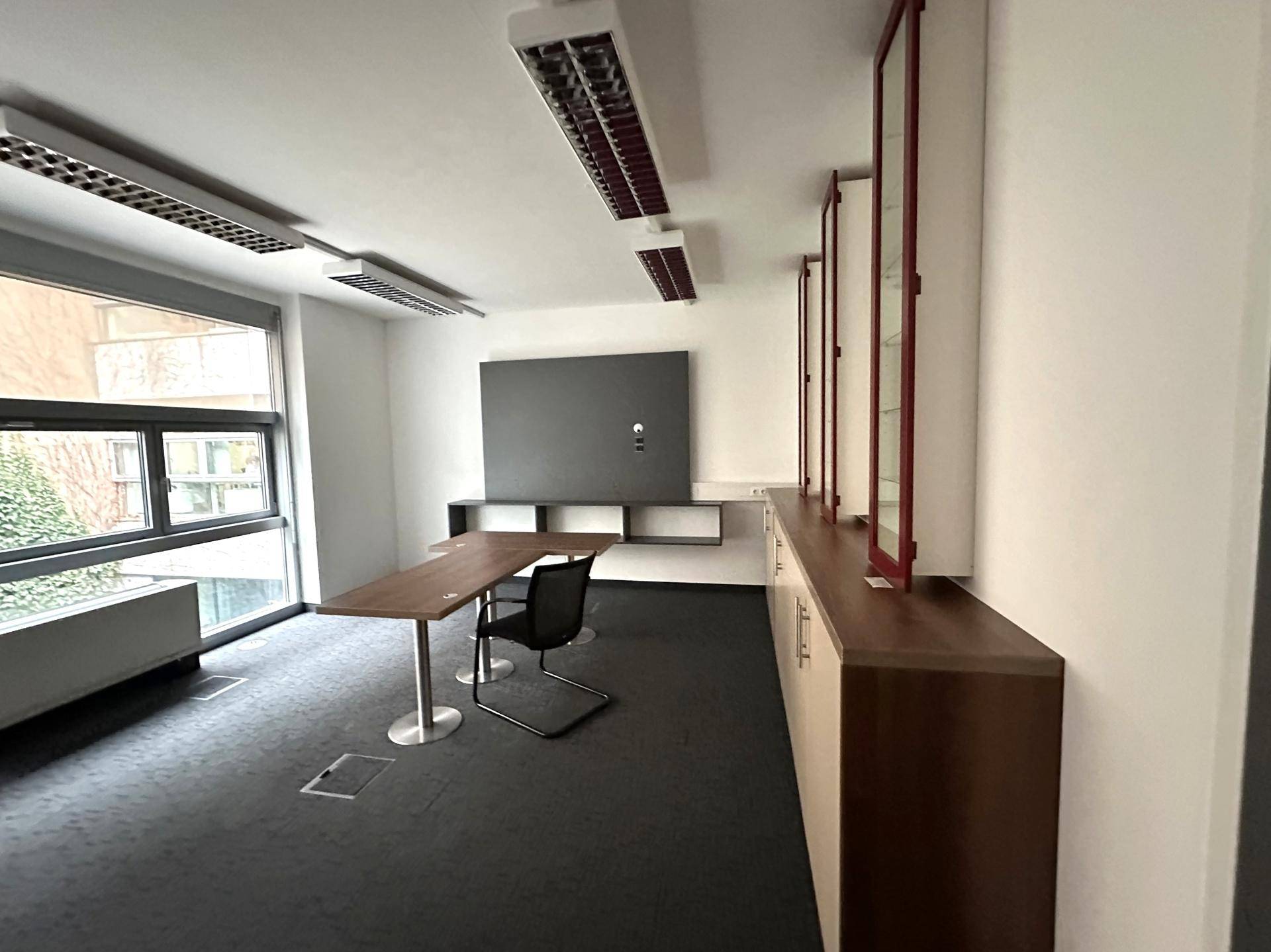 Modernes Büro mit Teppichboden, großem Fenster und funktionalen Möbeln, bereit zur Nutzung.